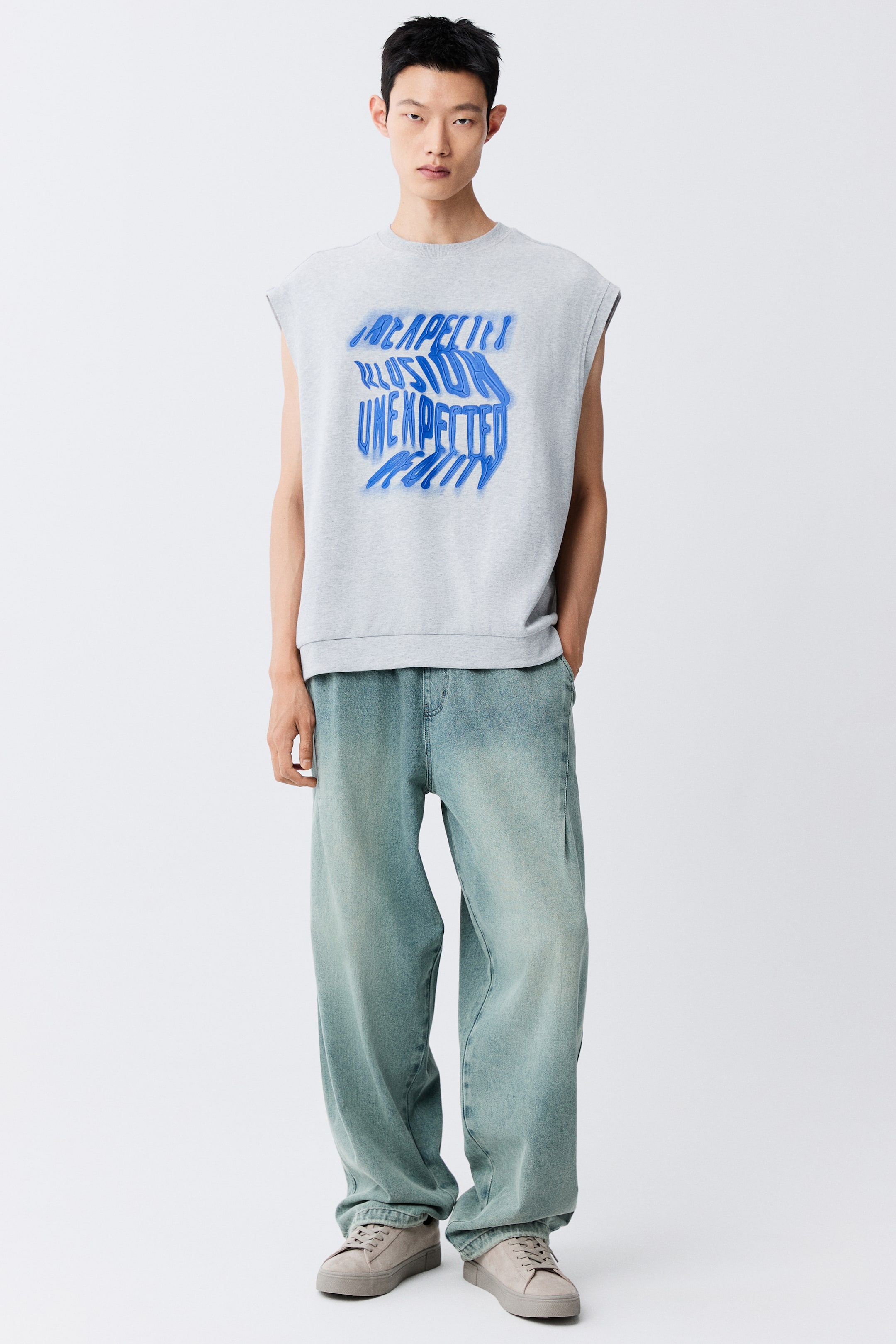 View larger image: The model is wearing jeans in a light greenish-blue denim wash, featuring a loose-fit balloon silhouette with forward-facing seams. The model is standing with one hand resting inside a diagonal side pocket, styling the jeans with a sleeveless grey graphic sweatshirt and light grey sneakers.