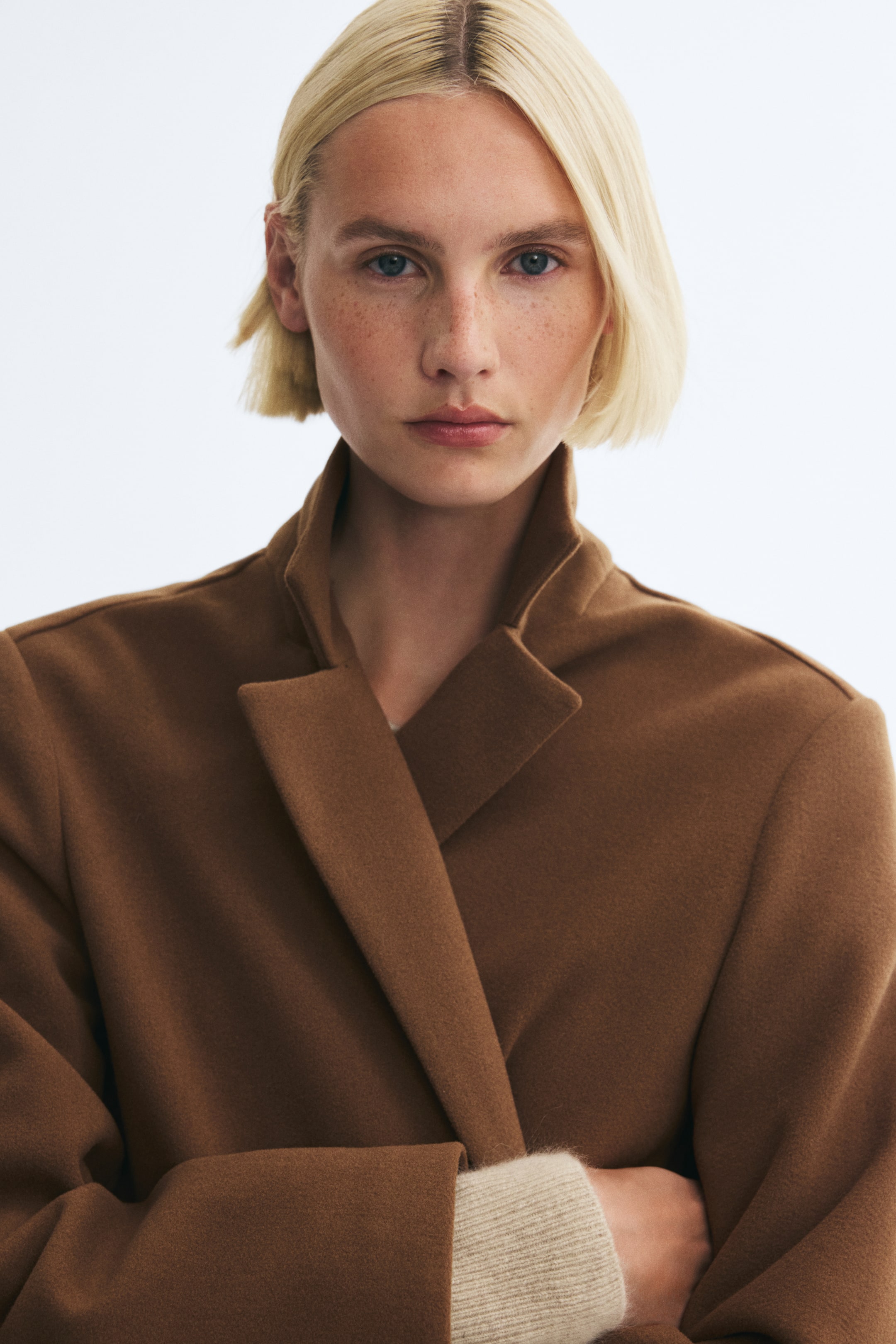 Voir une image plus grande: Le mannequin porte un manteau brun à simple boutonnage en tissu feutré doux, doté de larges revers crantés et d’un col relevé. Une manche en tricot côtelé beige pâle est visible sous le revers de la manchette du manteau, tandis que le mannequin se tient les bras croisés.