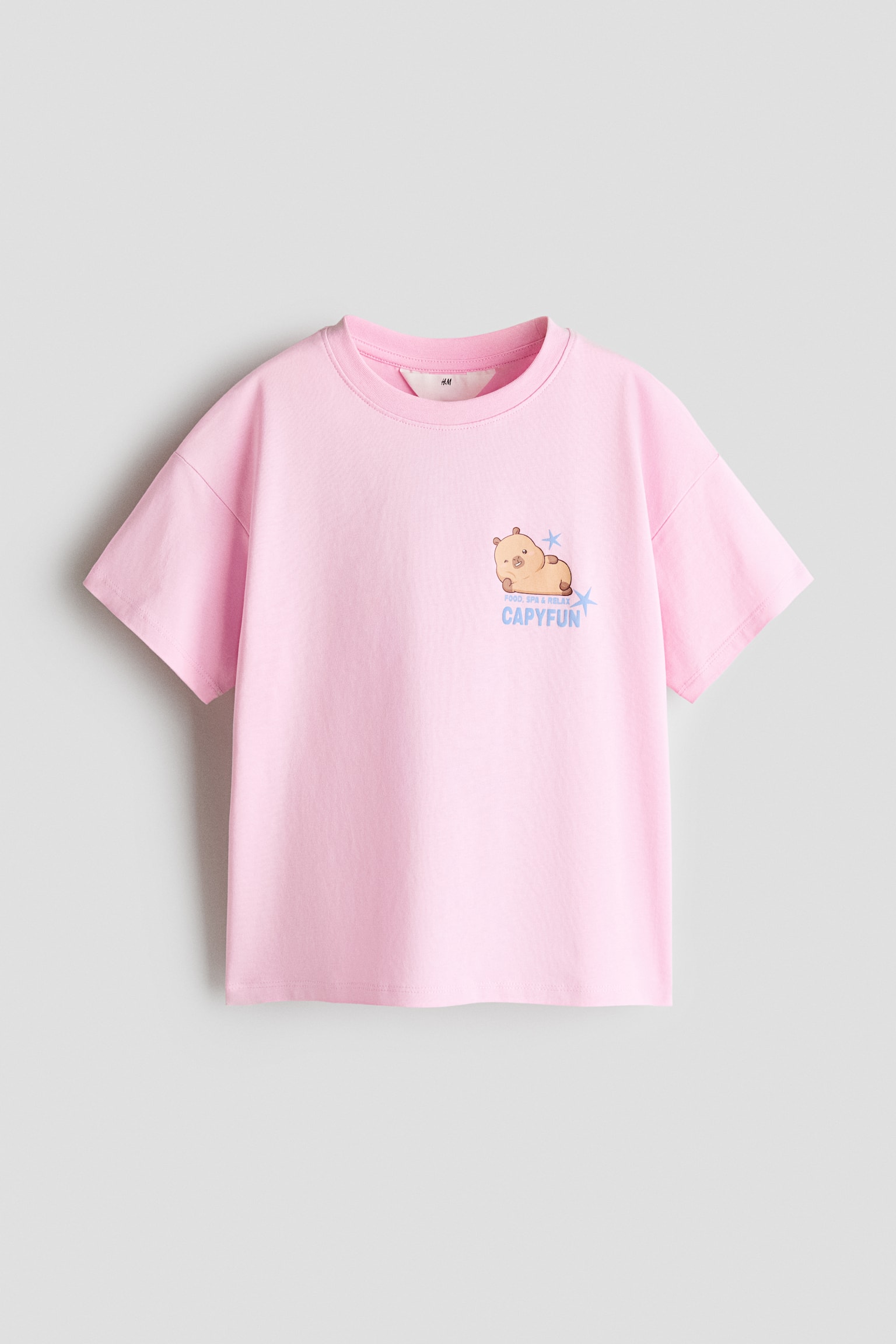 A light pink crew-neck T-shirt, shown from the front, displays a small graphic on the left chest. The graphic features a cartoon capybara lying down, with the blue text âCAPYFUNâ printed underneath.