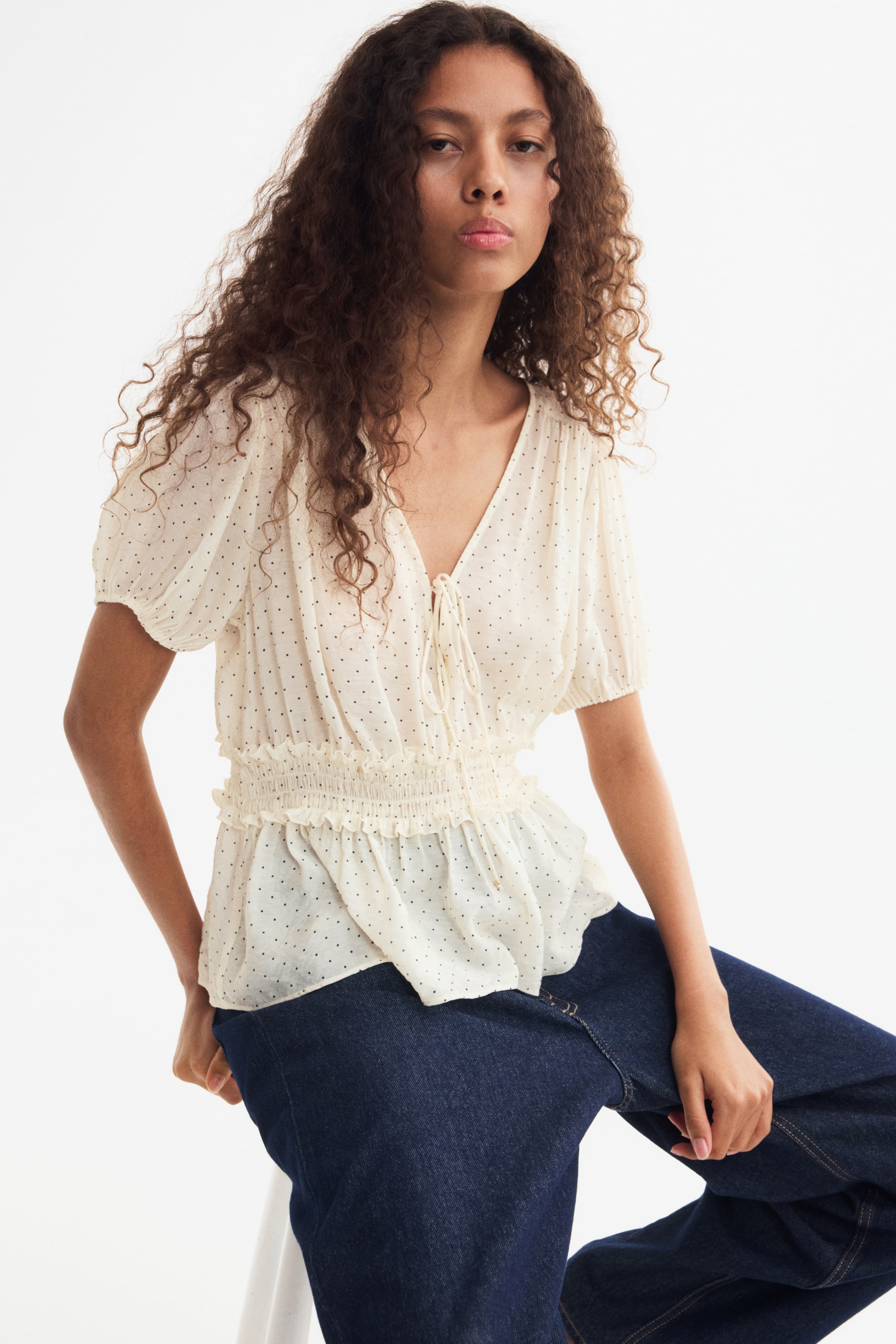 View larger image: A cream-coloured, dotted woven blouse is worn by the model, featuring a V-neckline with narrow front ties, short puff sleeves with elasticated cuffs, and a frilled smocked waist that flares into a peplum. The blouse is paired with dark blue jeans.