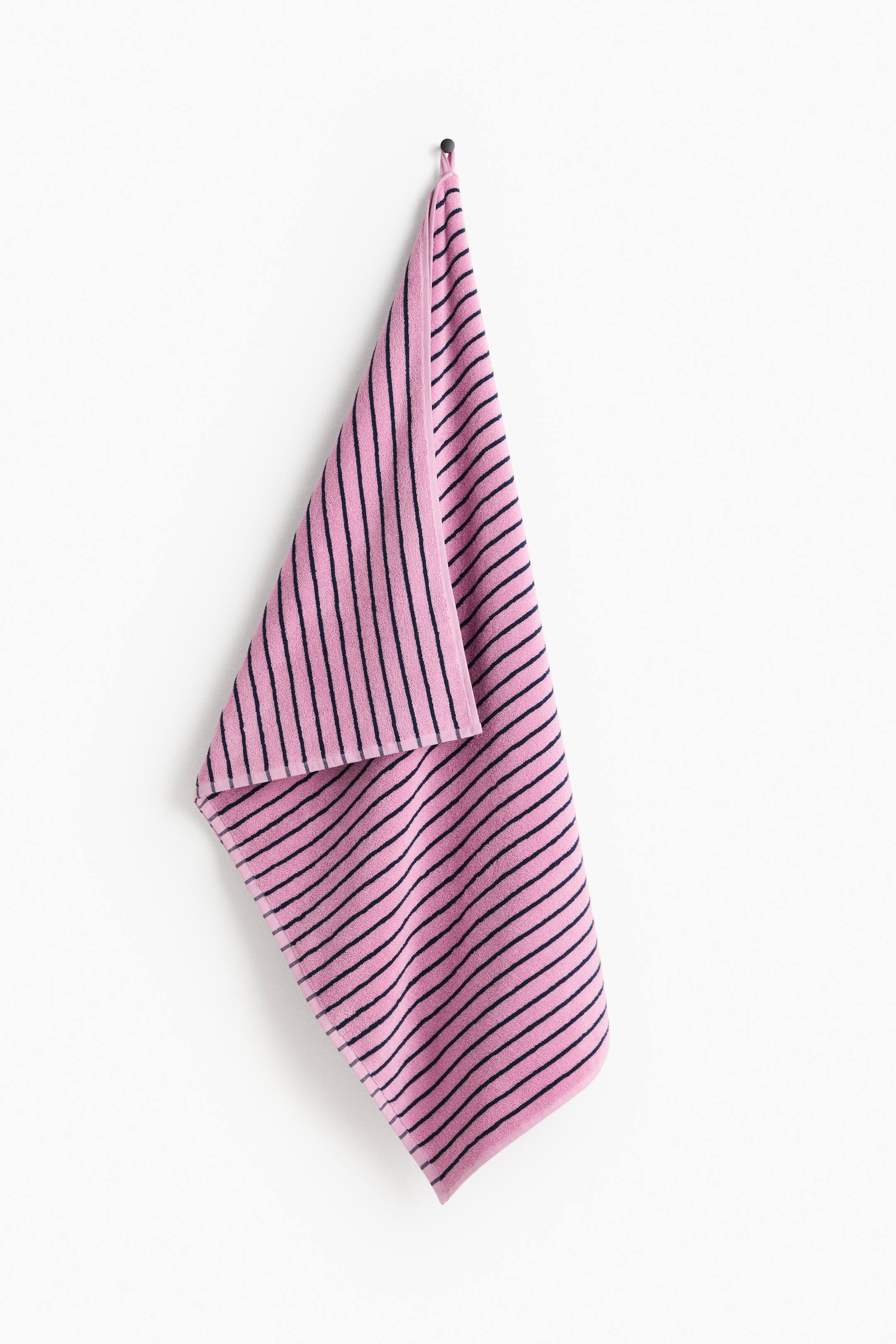 View larger image: A pink cotton terry towel featuring a dark navy blue striped pattern is hanging by its integrated loop from a small wall hook. The towel drapes naturally, showcasing its soft texture and striped design on both sides.