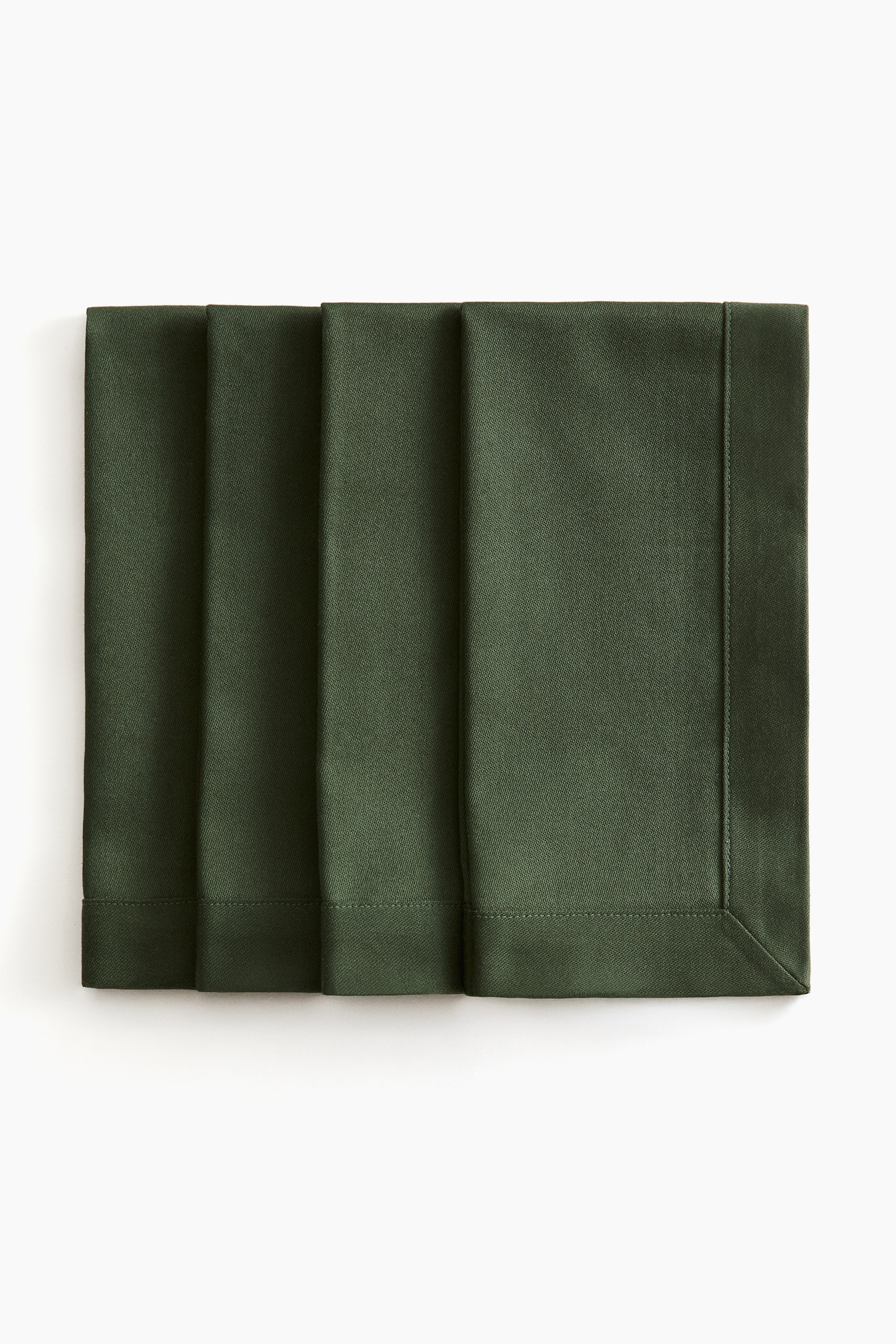 View larger image: Four dark green cotton napkins are overlapping in a row from a front view, displaying their structured rectangular shapes, folded hems with topstitching, and a mitred corner on the front napkin.