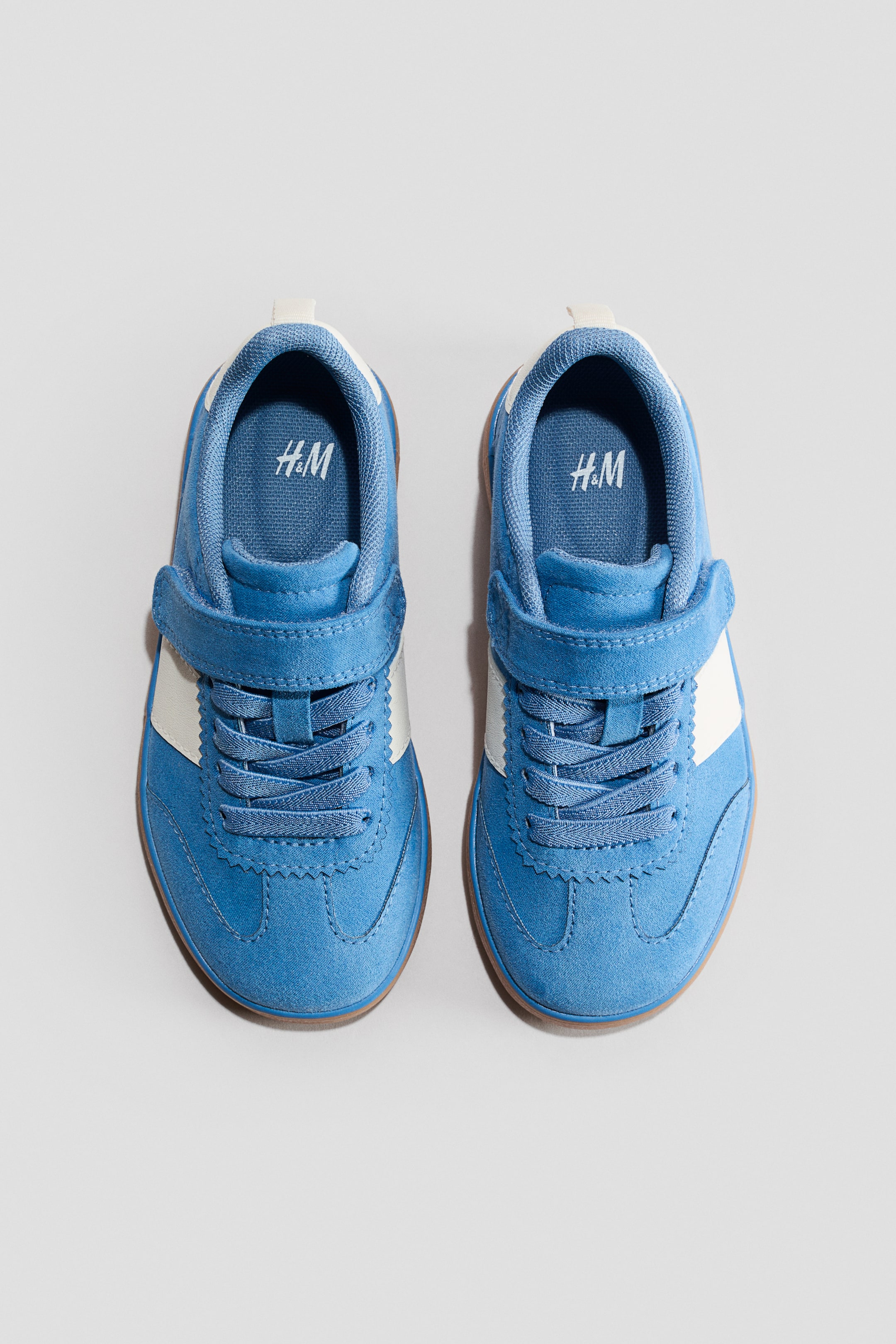 Grösseres Bild ansehen: Trainer. Ein Paar blaue Trainer wird von vorne gezeigt, mit weißen Seitenstreifen, gewellter Naht an den Zehen- und Ösenbereichen, einem Klettverschluss über den Schnürsenkeln und braunen Gummisohlen. H&M-Branding ist auf den Innensohlen sichtbar.