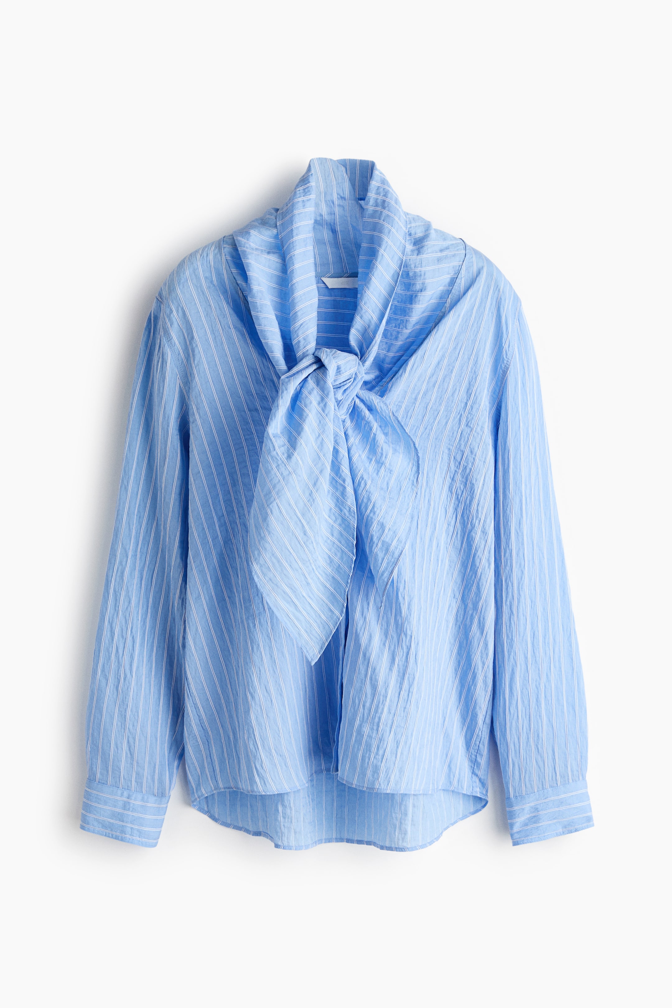View larger image: A light blue striped shirt is facing front, showcasing a crinkled texture and fine white vertical stripes. The shirt features a tie-neck detail tied in a bow, with long sleeves ending in buttoned cuffs and a subtly curved hem.