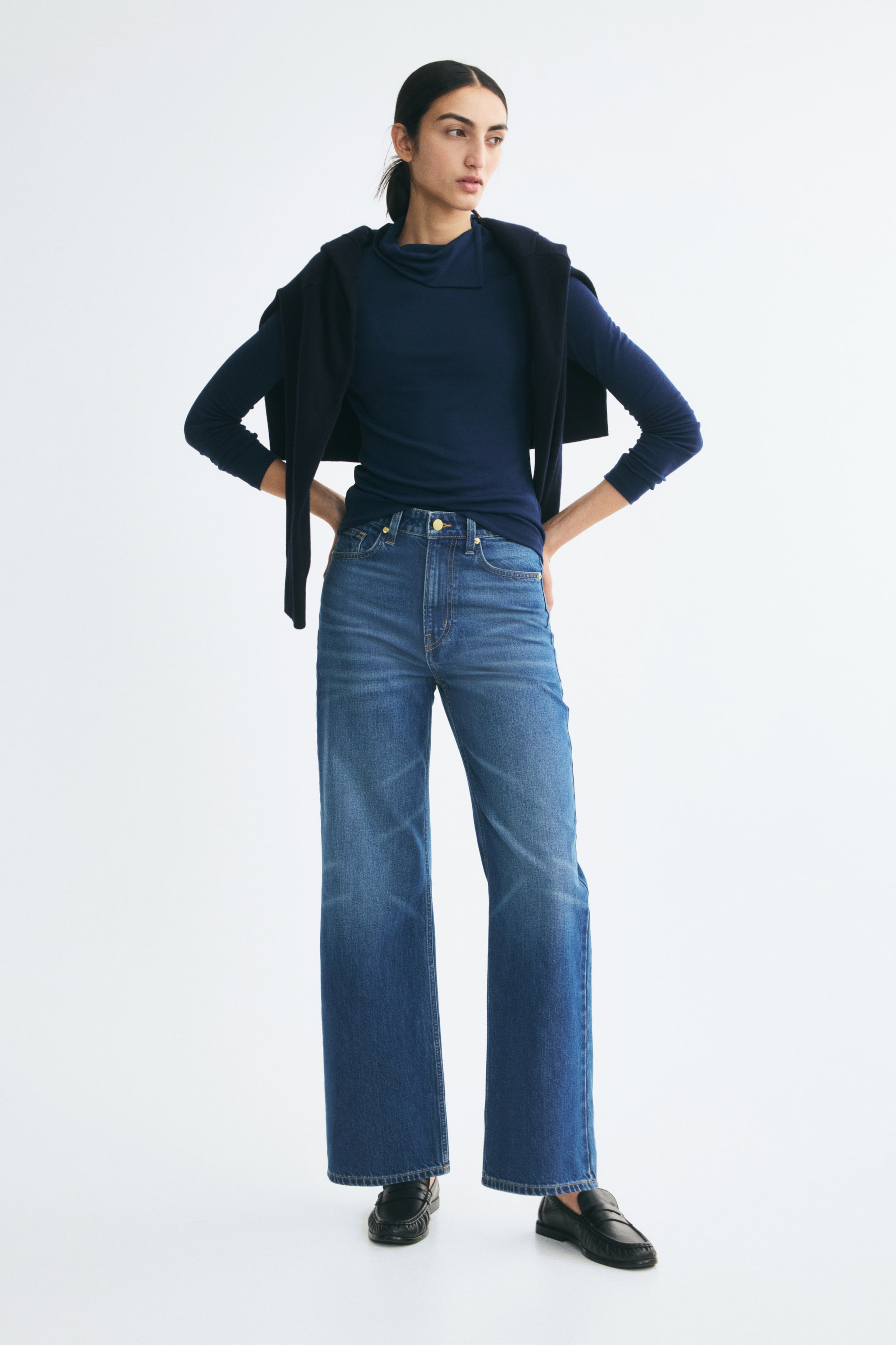 View larger image: Blue, high-waisted, straight-leg denim jeans are worn by the model, who is standing with hands on hips. The jeans feature a classic five-pocket design with light whiskering on the thighs and are styled with a tucked-in dark blue long-sleeve top and black loafers.