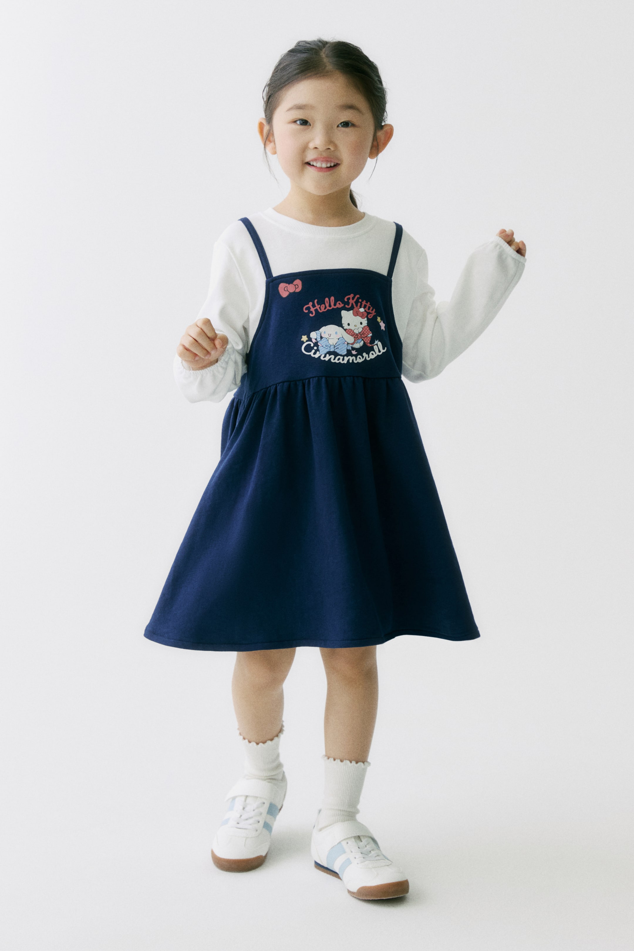拡大画像を表示: 正面からのアングルでネイビーのピナフォアドレスを撮影。細いスパゲッティストラップ、ウエストにギャザーの入ったAラインスカート、胸元に刺繍された「Hello Kitty Cinnamoroll」グラフィックが特徴で、白い長袖トップスの上に重ねています。