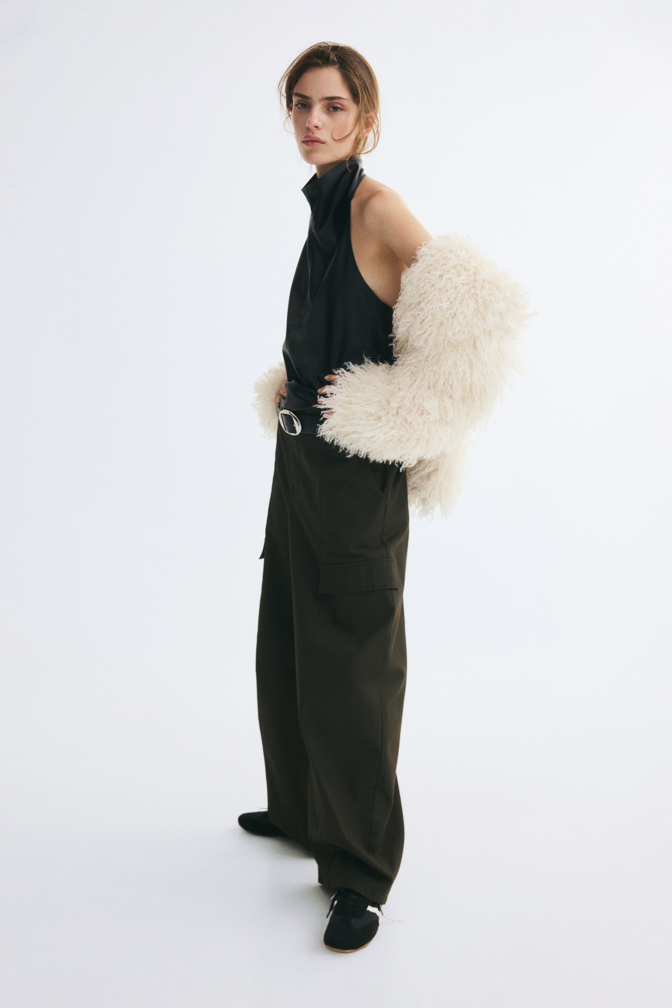 Größeres Bild anzeigen: Ein ärmelloses Neckholder-Top aus schwarzem beschichtetem Stoff wird vom Model getragen und in eine dunkle, weit geschnittene Hose gesteckt. Das Top ist mit einer flauschigen, strukturierten Jacke in Creme kombiniert, die lässig über die Arme des Models gelegt ist.