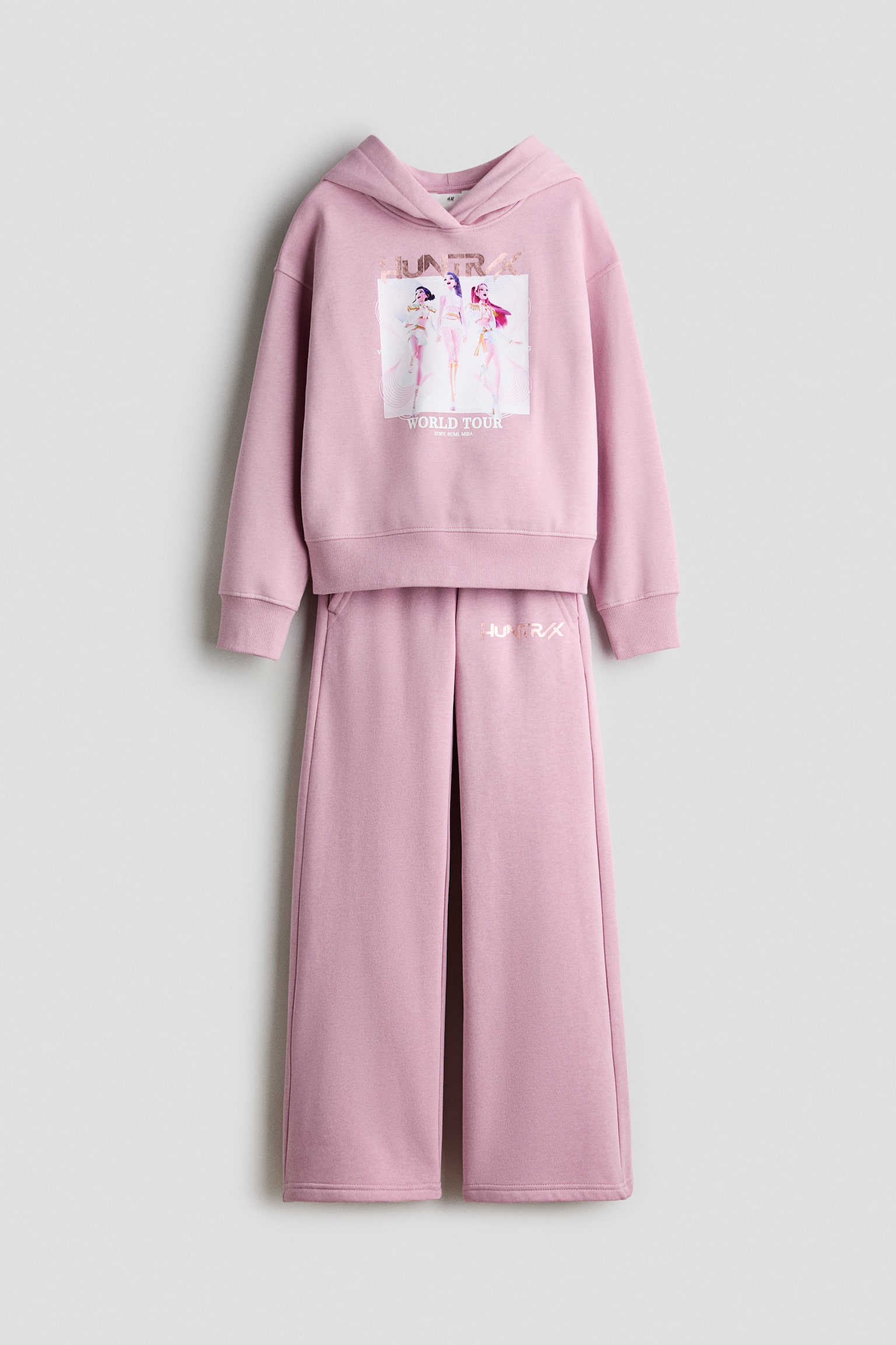 Ett set med rosa hoodie i borstad fleece och trÃ¤ningsbyxor visas framifrÃ¥n. Hoodien har ett grafiskt karaktÃ¤rstryck pÃ¥ brÃ¶stet och Ã¤r designad med huva med omlottsÃ¶ppning, nedhasade axlar och ribbade muddar, medan de matchande raka trÃ¤ningsbyxorna har resÃ¥r i midjan och en liten metallisk logotyp pÃ¥ Ã¶vre vÃ¤nstra lÃ¥ret.