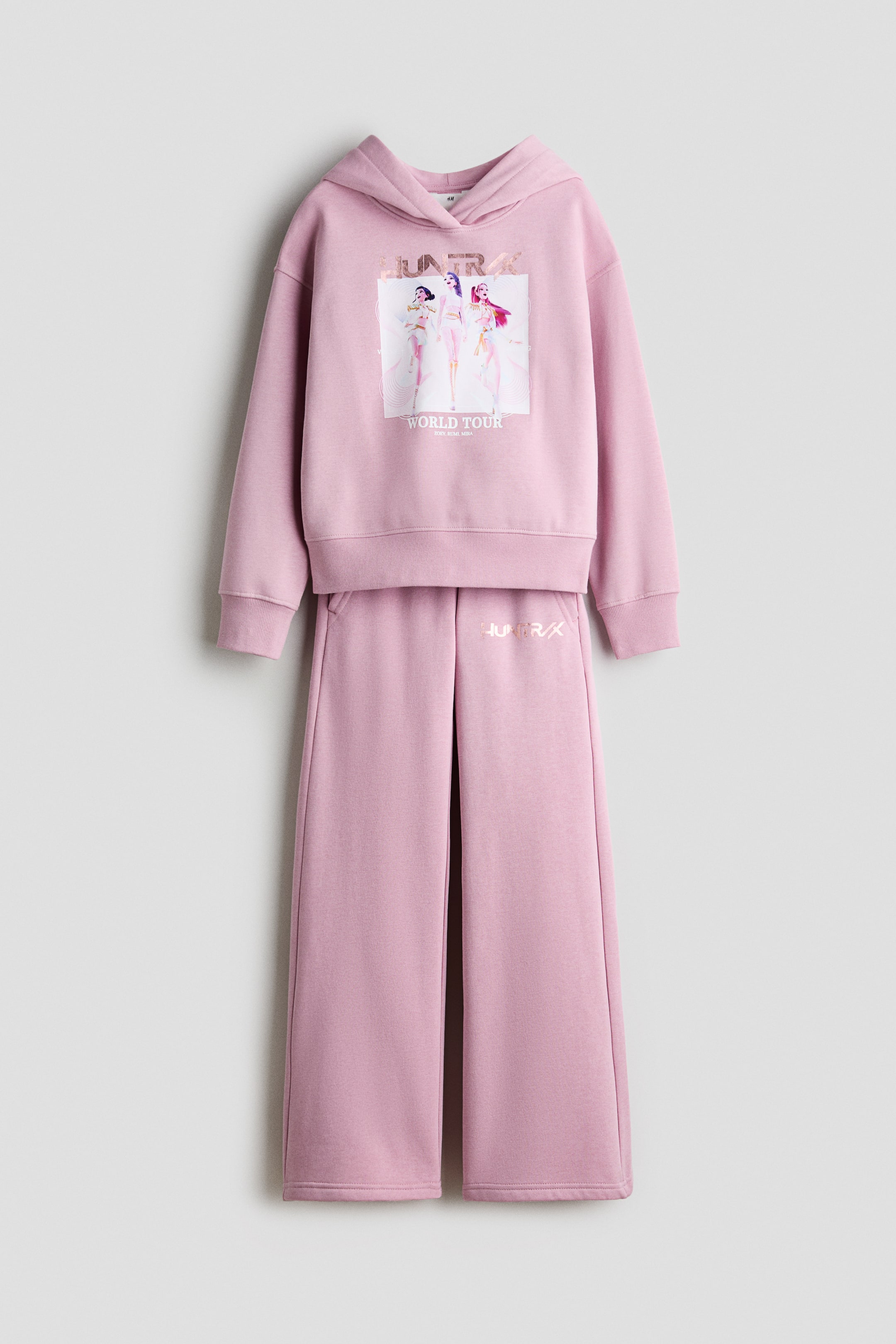 Grotere afbeelding bekijken: Een roze set van geborstelde fleece met hoodie en trainingsbroek wordt van voren getoond. De hoodie heeft een grafische karakterprint op de borst en is voorzien van een omslagkap, verlaagde schoudernaden en geribde boorden, terwijl de bijpassende straight-leg trainingsbroek een elastische tailleband en een klein metalen logo op de linker bovenbeen heeft.
