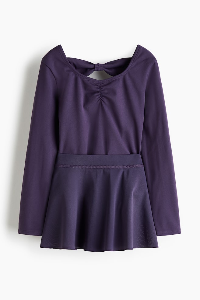 A dark purple long-sleeved leotard with an attached skirt is lying flat, showcasing its back view. The leotard is featuring a wide open back with a tied bow at the top and ruching below, and the attached skirt is flaring out from a defined waistband. - 1