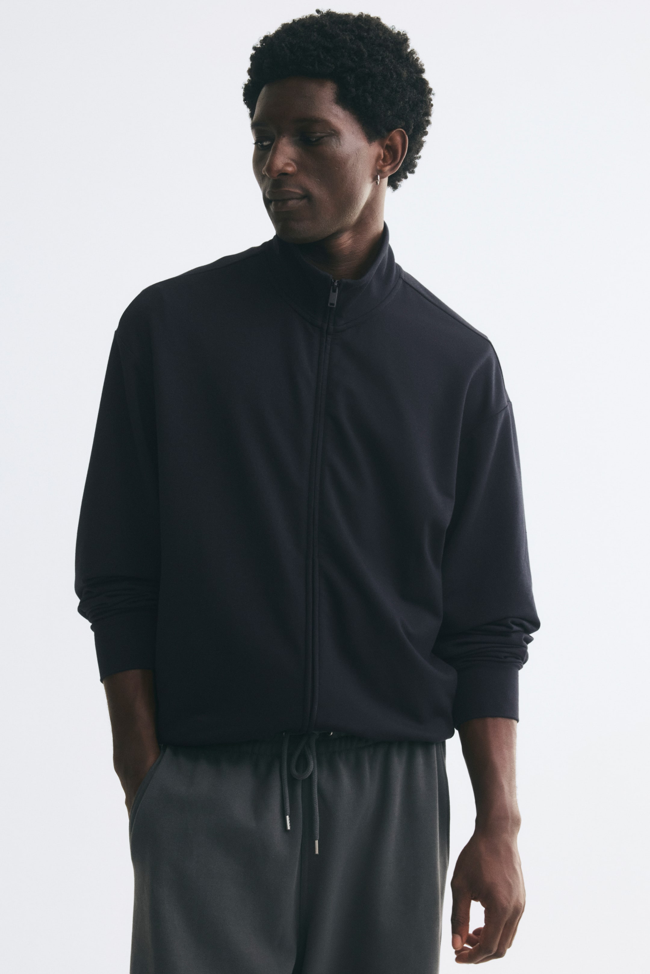 Voir une image plus grande: Une veste en tissu molletonné noir avec col montant, fermeture éclair à l’avant et épaules tombantes est portée par le mannequin. La veste coupe décontractée est agencée avec un pantalon à cordon de serrage gris foncé.