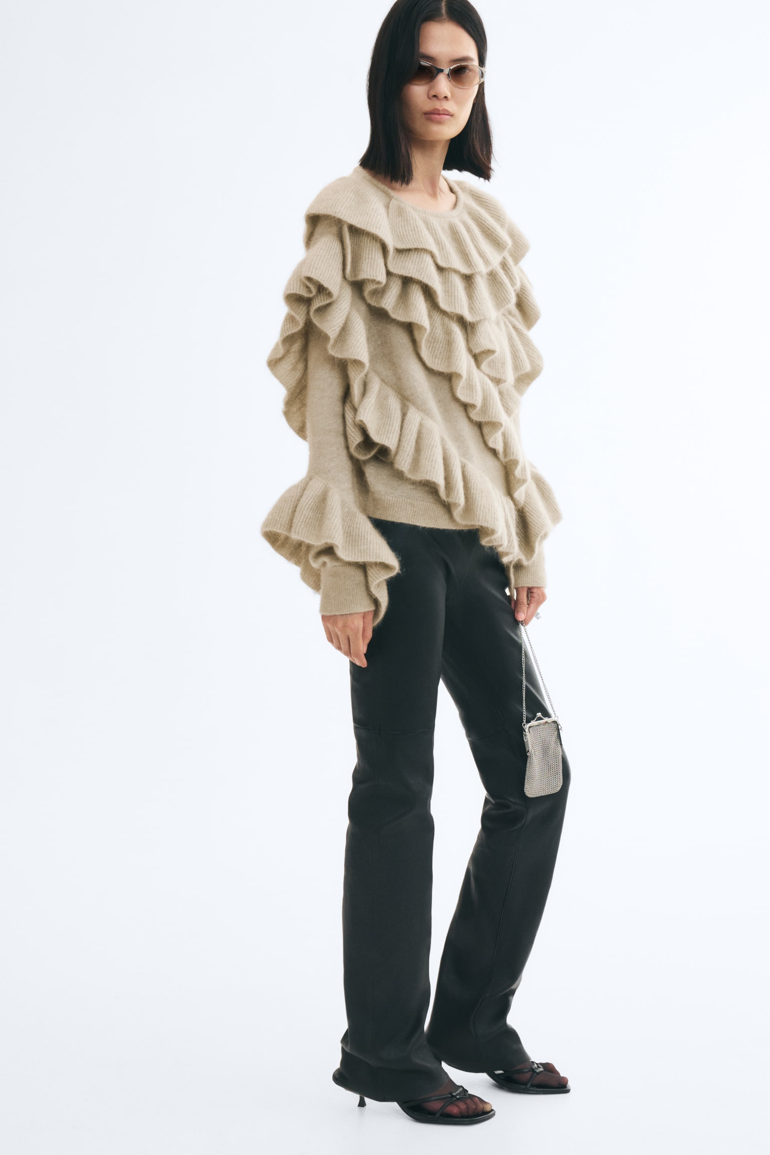A beige knit jumper featuring tiered ribbed flounces throughout the front and sleeves is being worn by the model. The jumper has a round neckline, dropped shoulders, and long sleeves with flared ruffled cuffs, styled here with black trousers. - 1