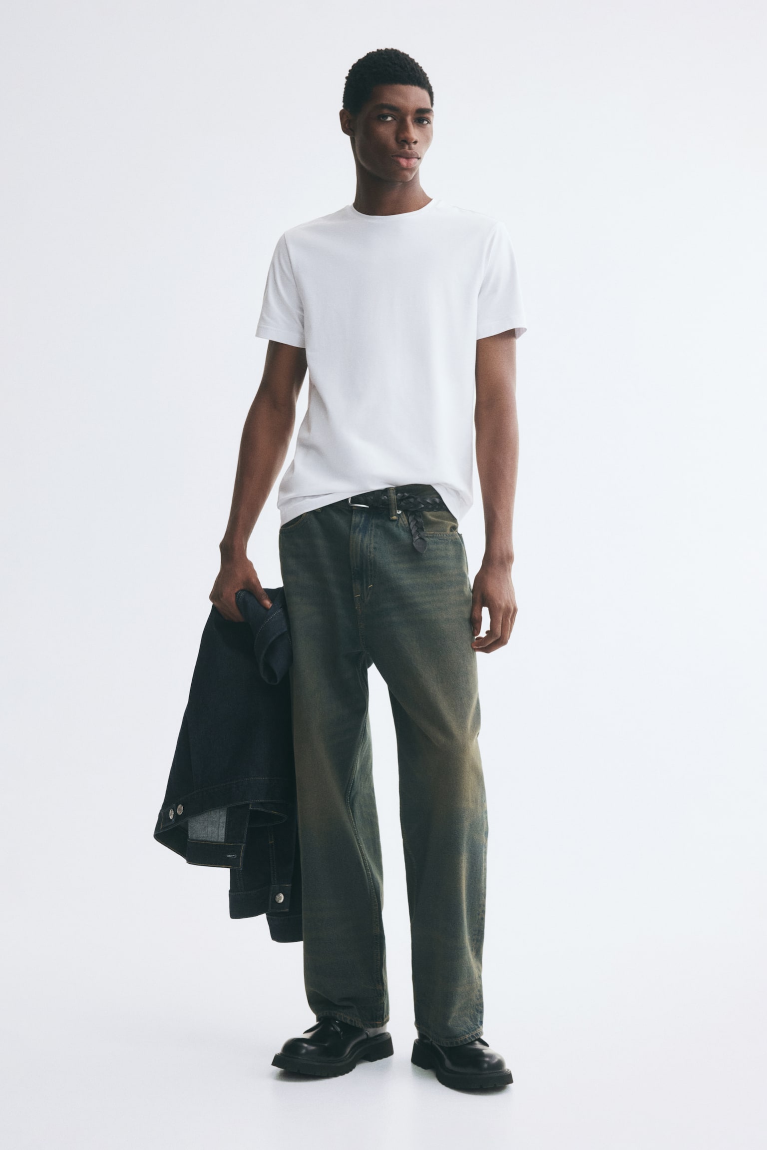 Wide-leg denim jeans in a faded, greenish-tinted wash are worn by the model, styled with a tucked-in white crew-neck T-shirt and a black braided belt. The model is also carrying a dark-wash denim jacket in one hand. - 1