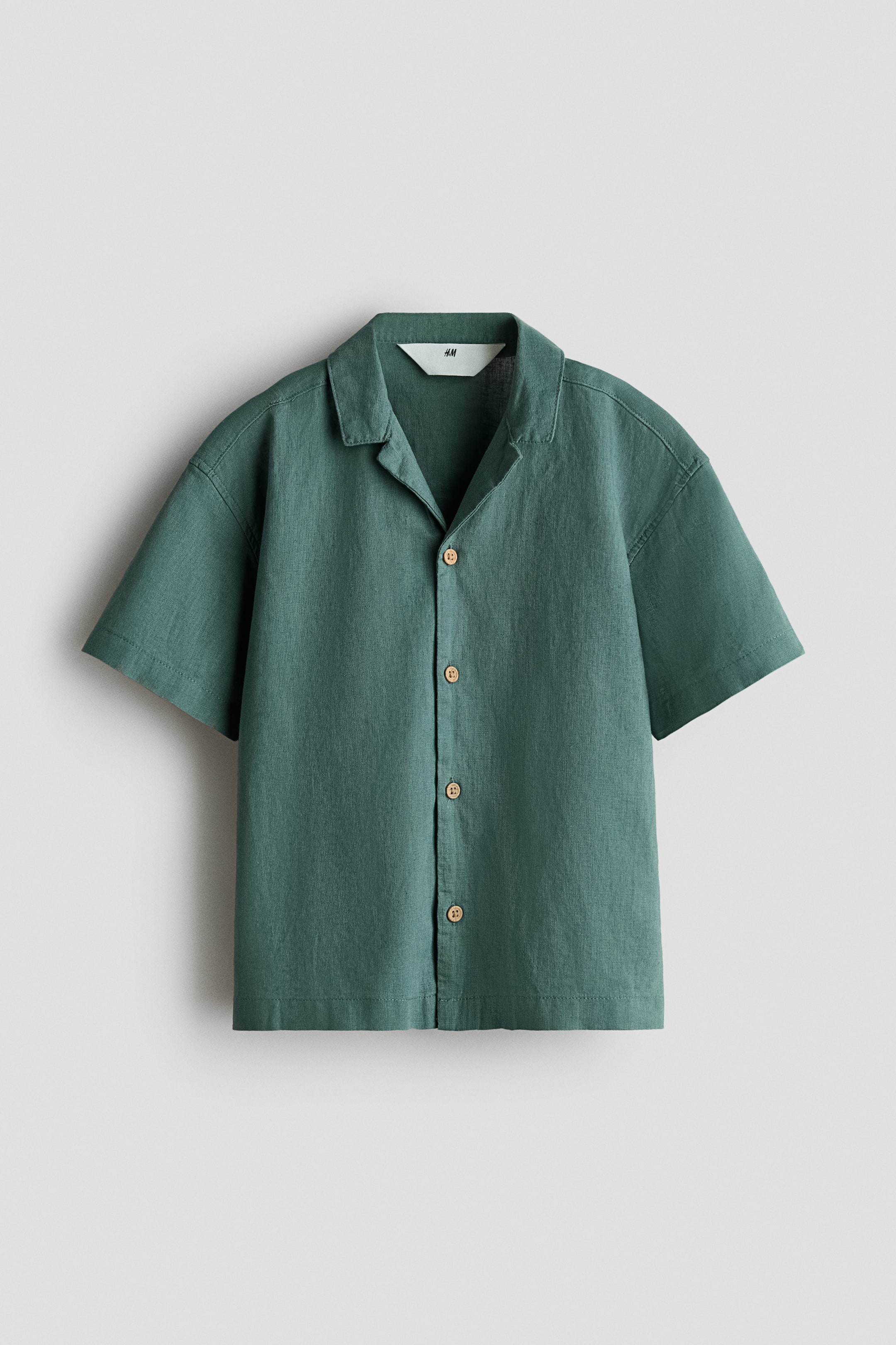 View larger image: A short-sleeved shirt is facing front, showcasing an airy dark green linen and cotton blend. It features a resort collar, gently dropped shoulders, and light brown buttons running down its front.