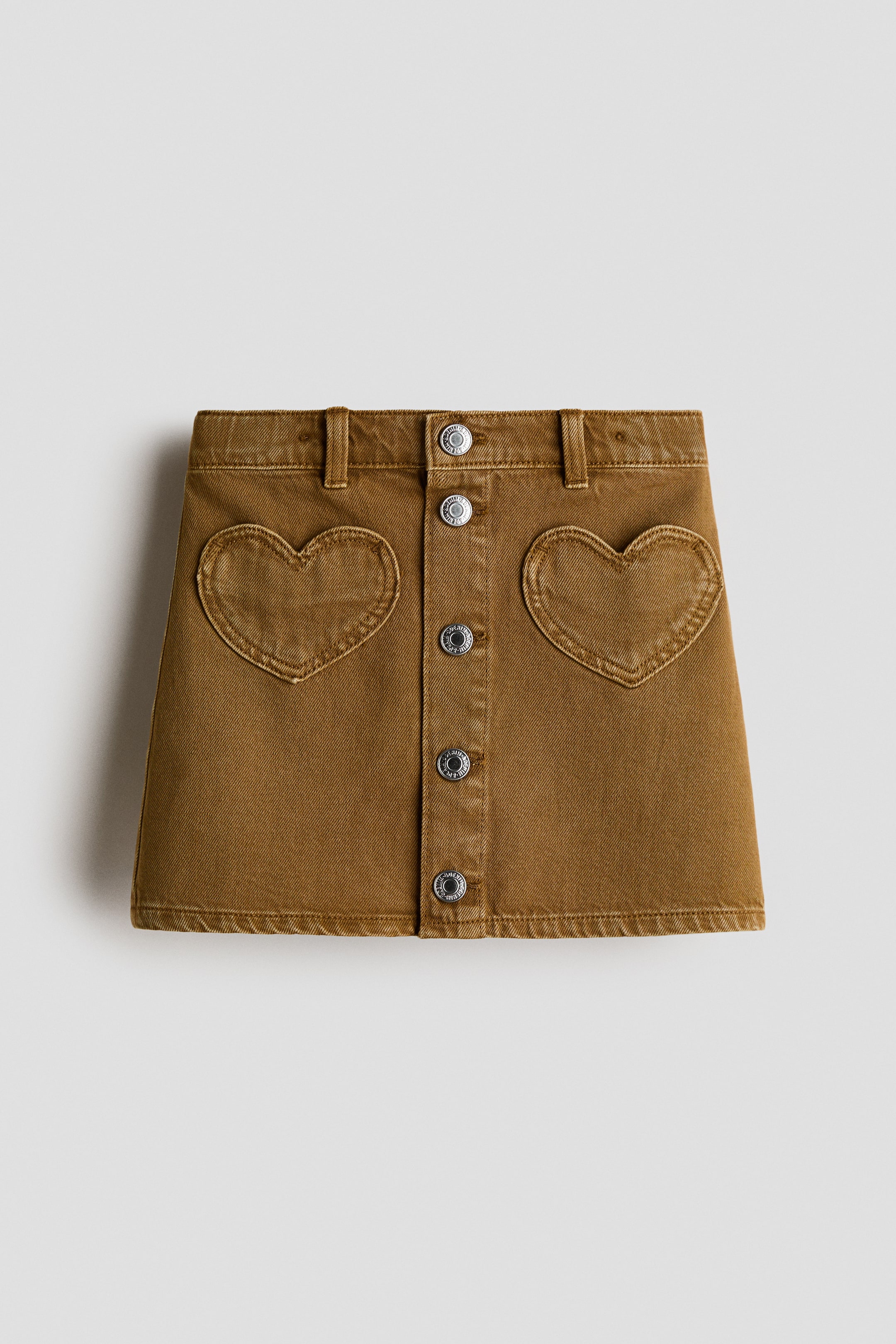 View larger image: A camel-coloured denim mini-skirt is showcasing its front, featuring a full button-front closure with six silver-toned metal buttons. It is presenting two heart-shaped patch pockets, one on each side of the button placket.