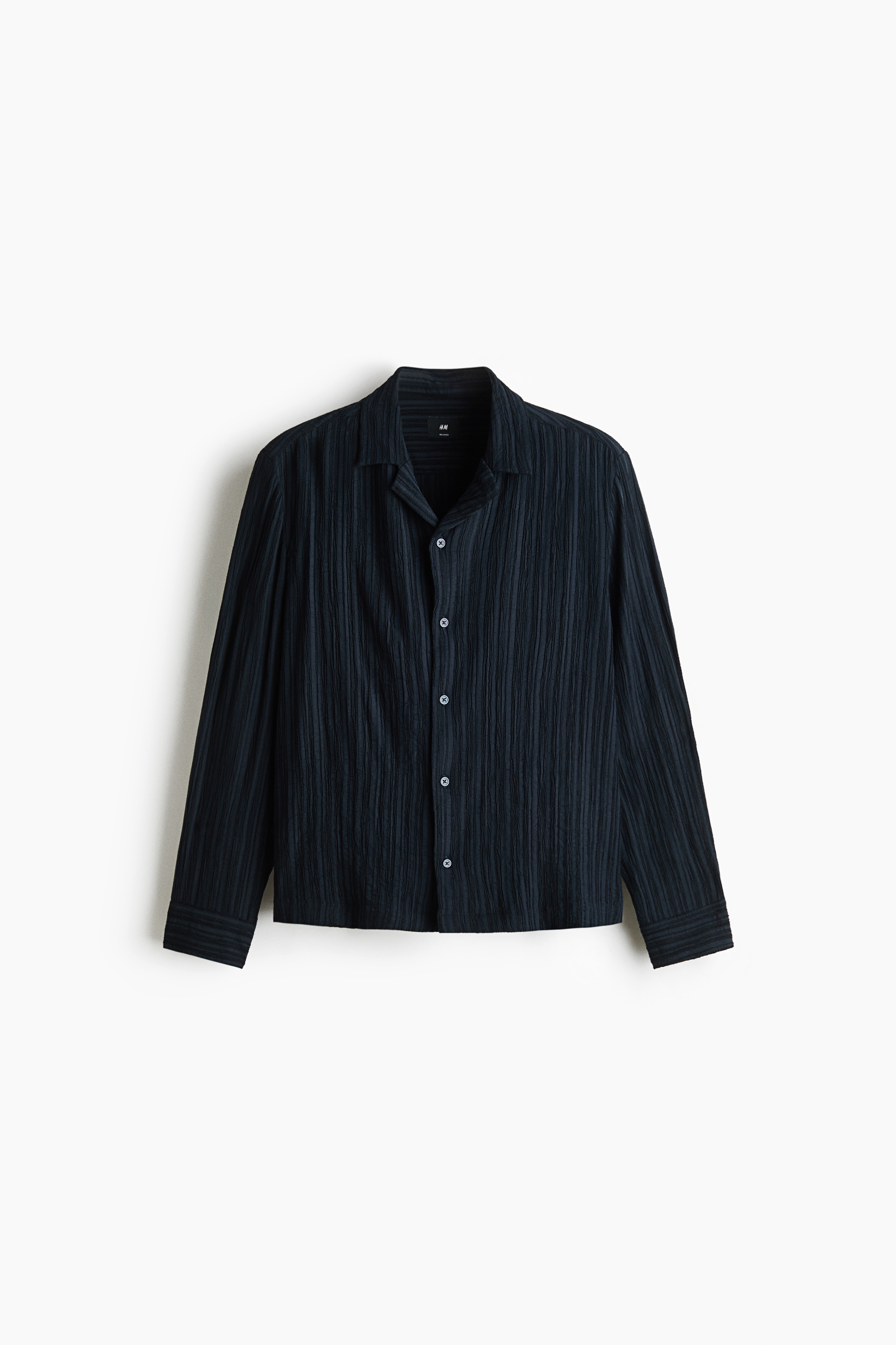 A dark navy, long-sleeved shirt in a textured crinkle weave is being worn by the model, with the front loosely tucked into blue jeans. The relaxed-fit shirt features a resort collar and a button-front closure.