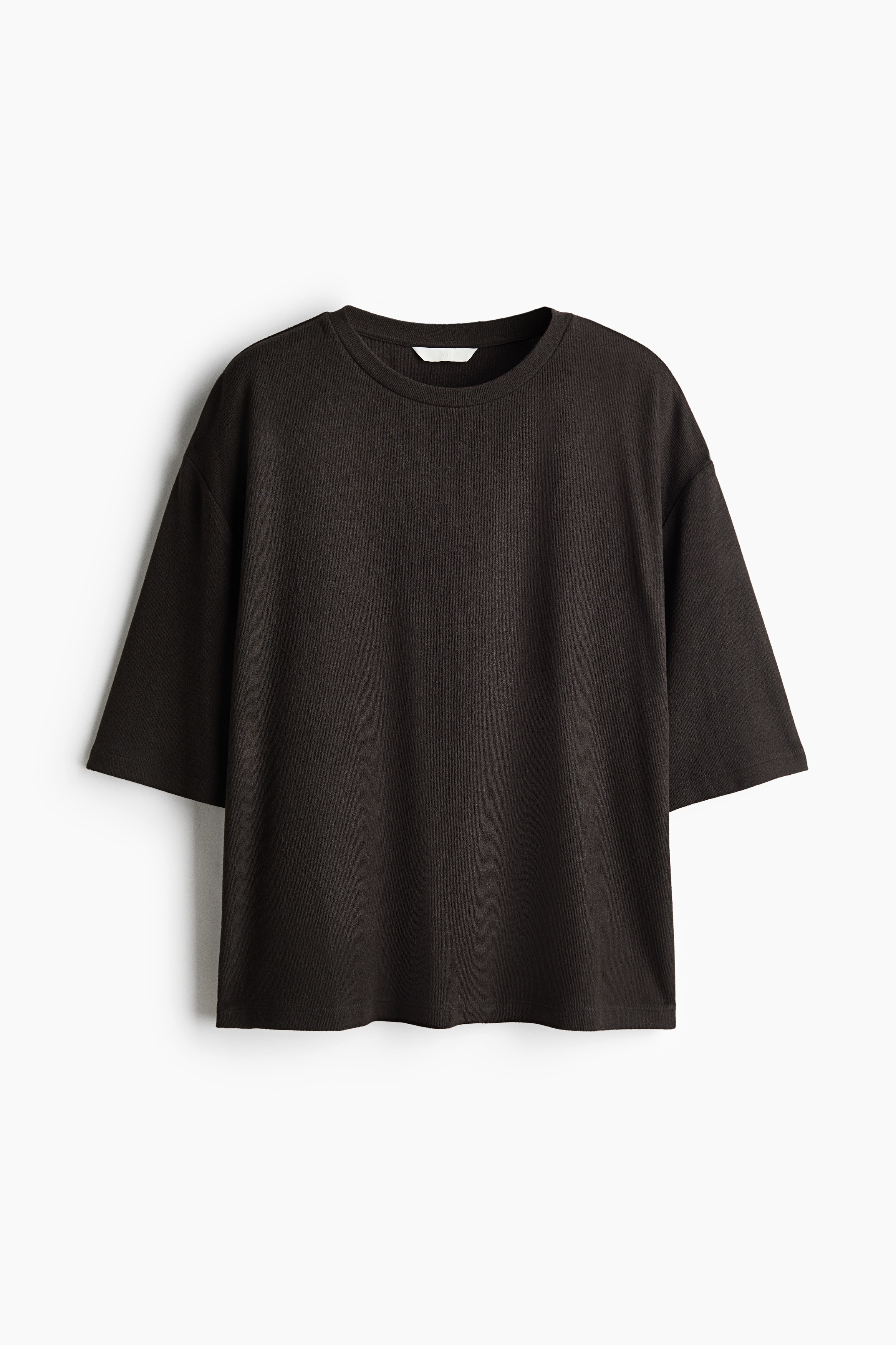 A dark brown, relaxed-fit crew neck T-shirt with elbow-length sleeves is worn by the model, who is standing with arms crossed. The T-shirt is styled tucked into light-wash, wide-leg blue jeans secured with a black belt.