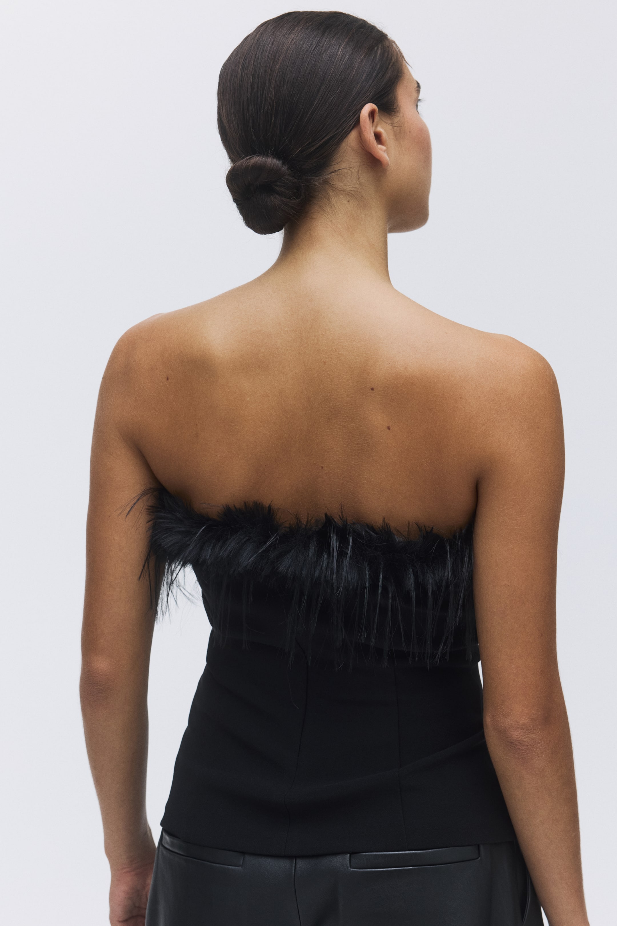 Agrandir l'image: Un haut noir sans bretelles, orné d'une bordure de plumes le long de son bord supérieur, est vu de l'arrière. Le haut ajusté est porté avec un pantalon noir.