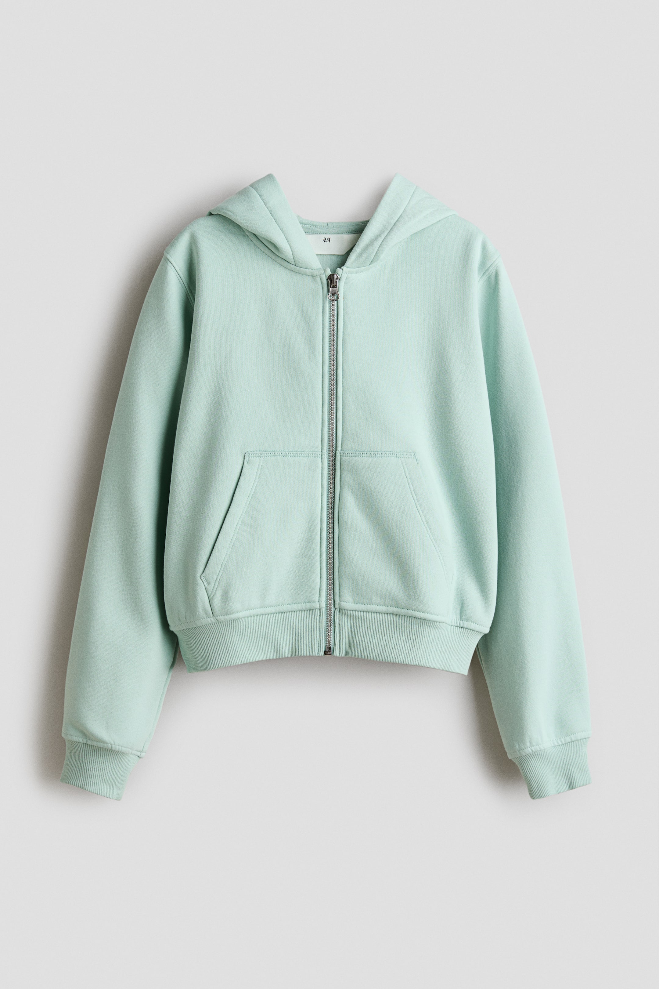 View larger image: A light green zip-through hoodie is showcasing its front, featuring a jersey-lined hood, a silver-tone zip running down the centre, two front patch pockets, ribbed cuffs, and a ribbed hem.