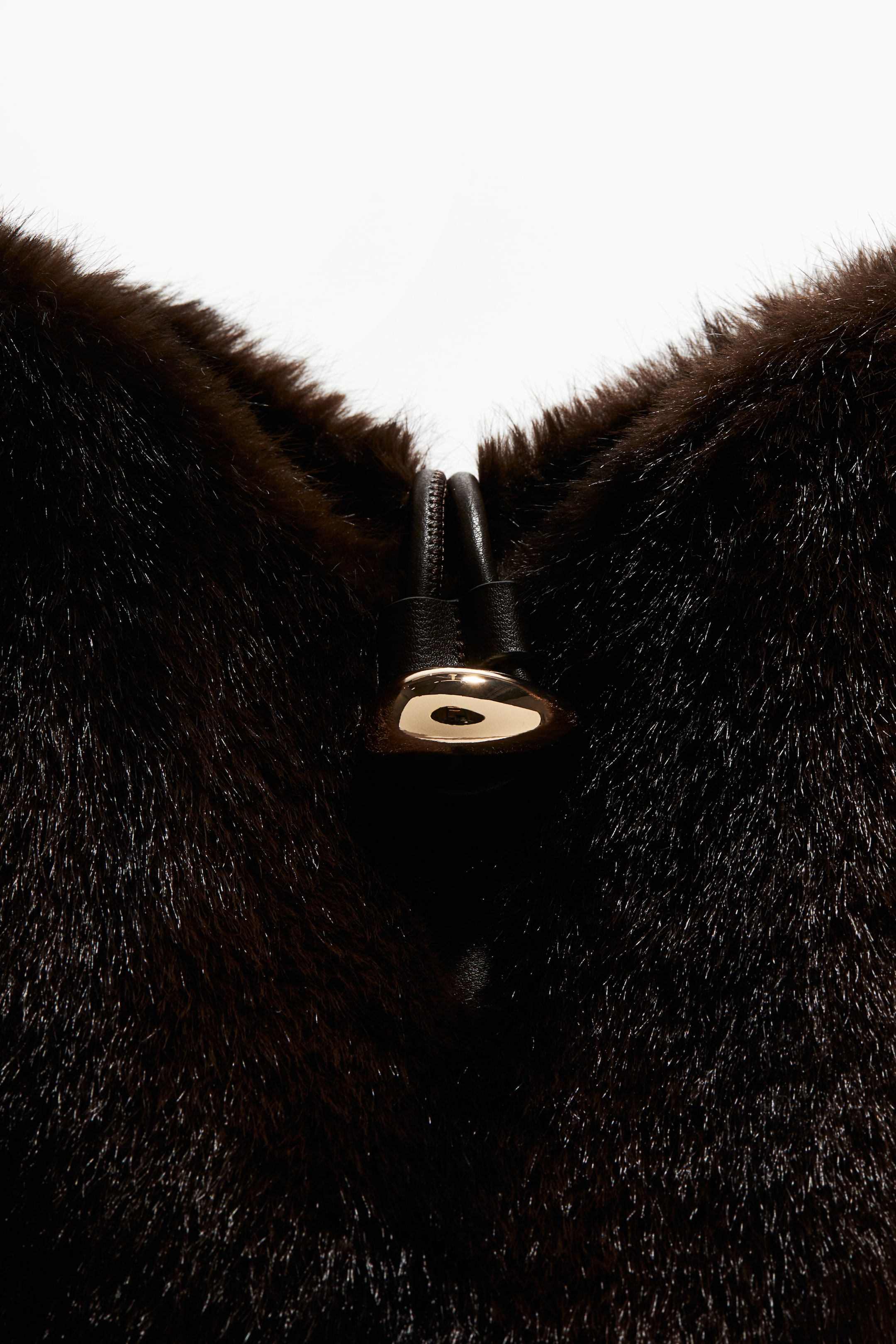 Ver imagen más grande: Un bolso de cubo marrón oscuro presenta un exterior suave, similar a la piel. Su apertura superior se cierra con un lazo negro que pasa a través de un detalle metálico de tono dorado en la PARTE DELANTERA.