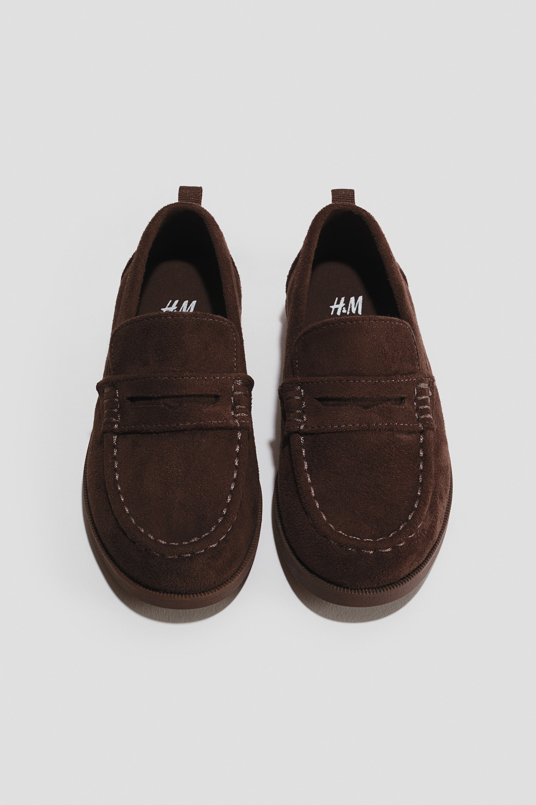 View larger image: A pair of dark brown suede loafers is shown from a top-down perspective, displaying their structure and design. The loafers feature a classic penny keeper strap across the vamp, contrast-stitched moccasin seams around the toe box, and small pull tabs at the heels.