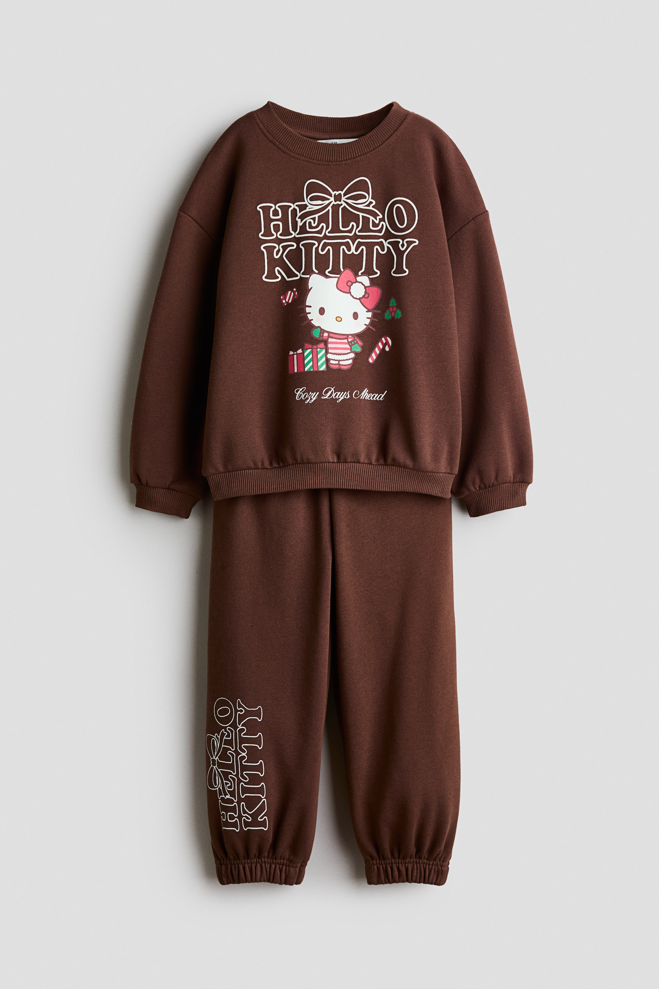 Ver imagem maior: É apresentado, visto de frente, um conjunto a condizer de camisola polar castanha e calças de fato de treino, com um gráfico natalício da Hello Kitty estampado no peito. A camisola tem decote redondo com acabamento canelado, ombros descaídos e punhos e bainha canelados, enquanto as calças de fato de treino a condizer apresentam bainhas elásticas e o texto vertical "Hello Kitty" estampado na parte inferior da perna direita.