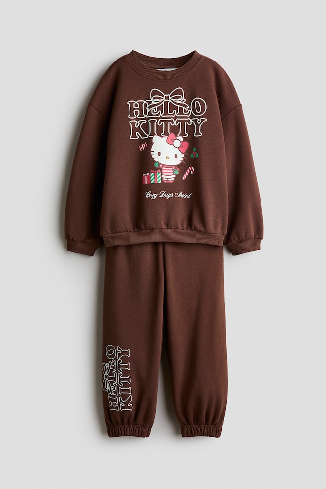 Ein passendes Set aus braunem Fleece-Sweatshirt und Trainingshose ist von vorne zu sehen und zeigt einen aufgedruckten Hello Kitty Holiday-Print auf der Brust. Das Sweatshirt verfÃ¼gt Ã¼ber einen runden, gerippten Ausschnitt, Ã¼berschnittene Schultern sowie gerippte BÃ¼ndchen und einen gerippten Saum, wÃ¤hrend die dazu passende Trainingshose elastische BeinabschlÃ¼sse und den vertikal aufgedruckten Schriftzug âHello Kittyâ am unteren rechten Bein aufweist. - 1