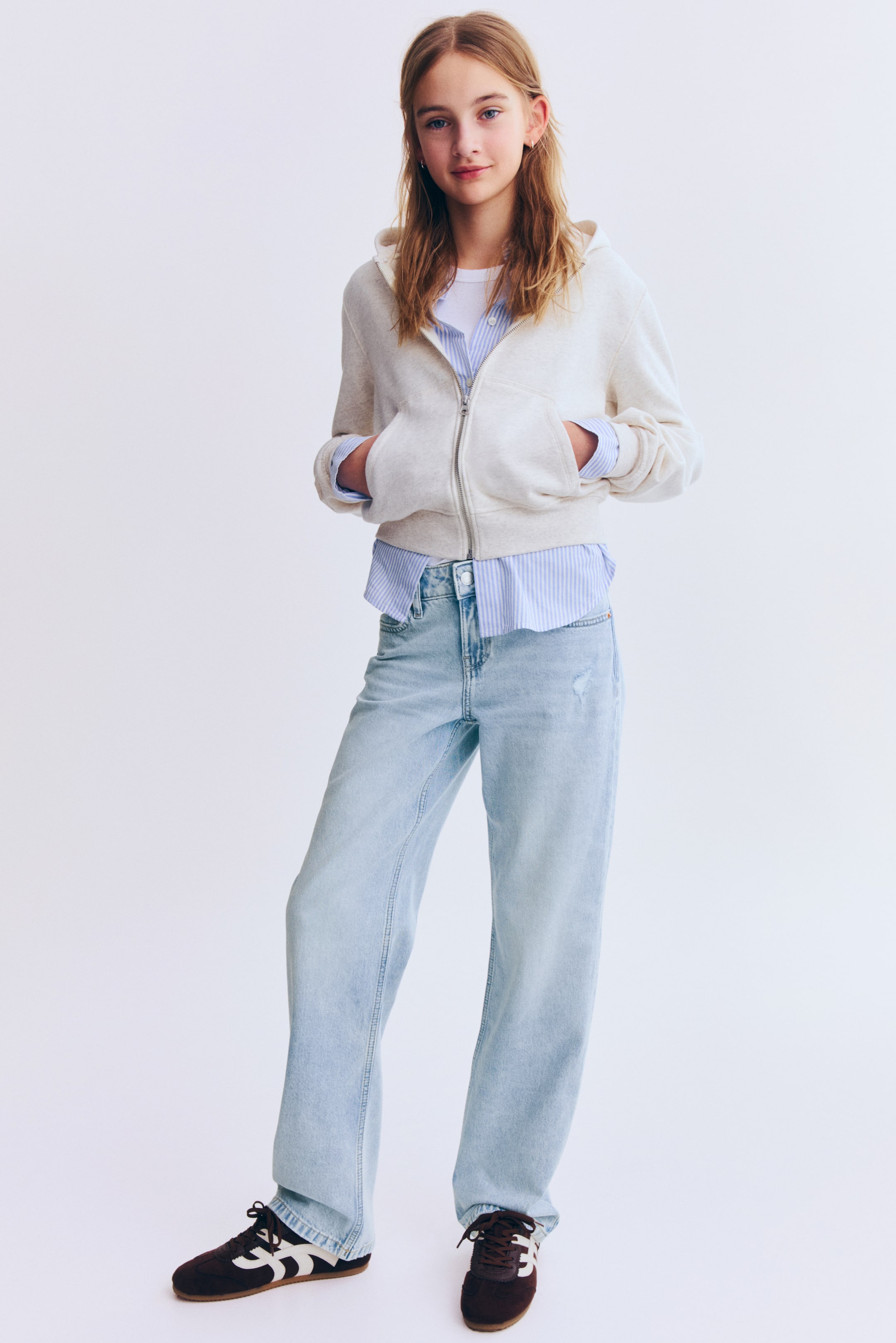 View larger image: A light beige full-zip hoodie with ribbed cuffs and hem is layered over a light blue and white striped button-up shirt and a white T-shirt. It’s paired with light-wash relaxed-fit jeans and dark brown suede sneakers with white stripes.