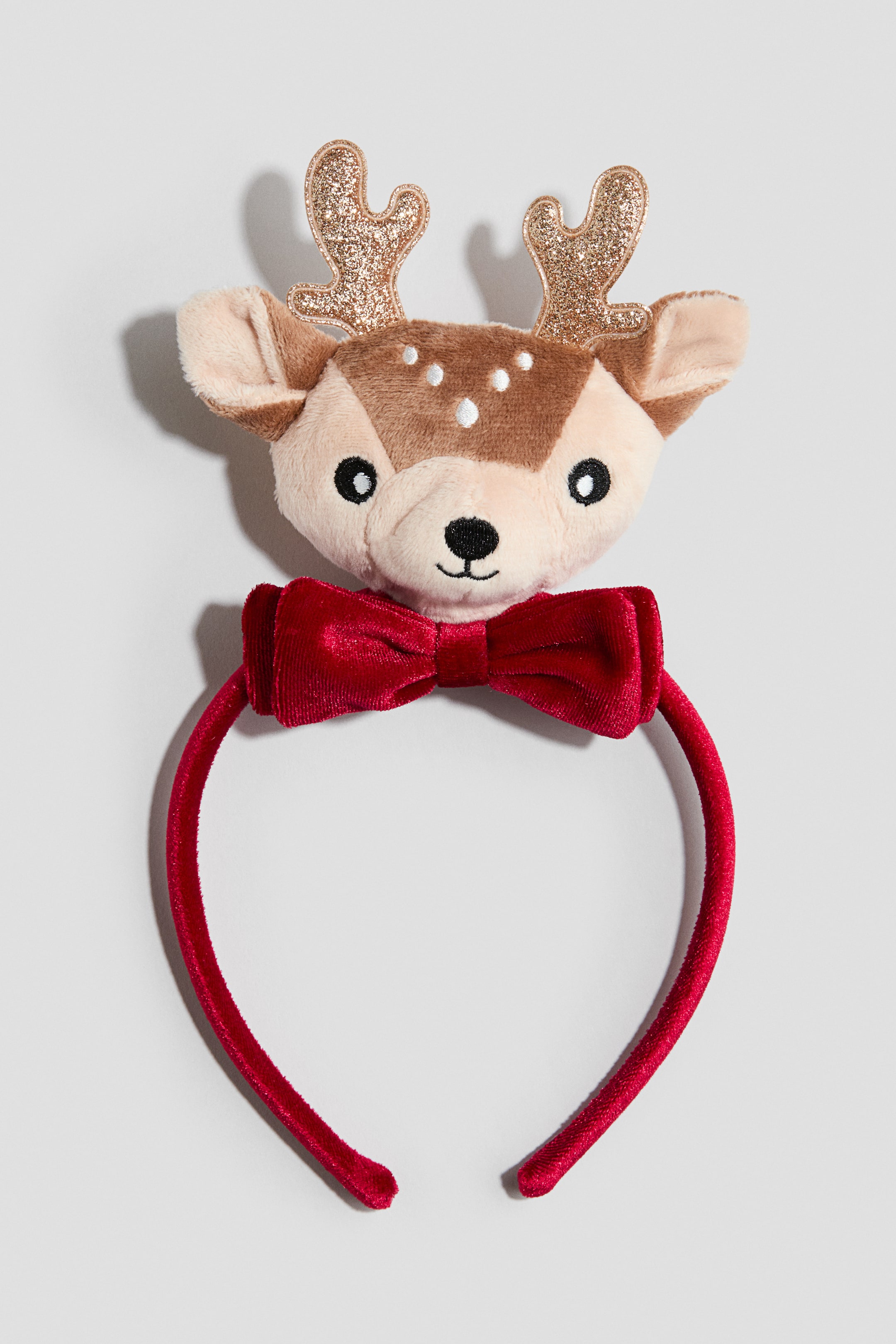 View larger image: A red velvet-covered headband features a plush deer head facing forward, adorned with gold glitter antlers and a red velvet bow tie underneath. The plush deer features embroidered black eyes and mouth, white spots on its forehead, and soft ears.