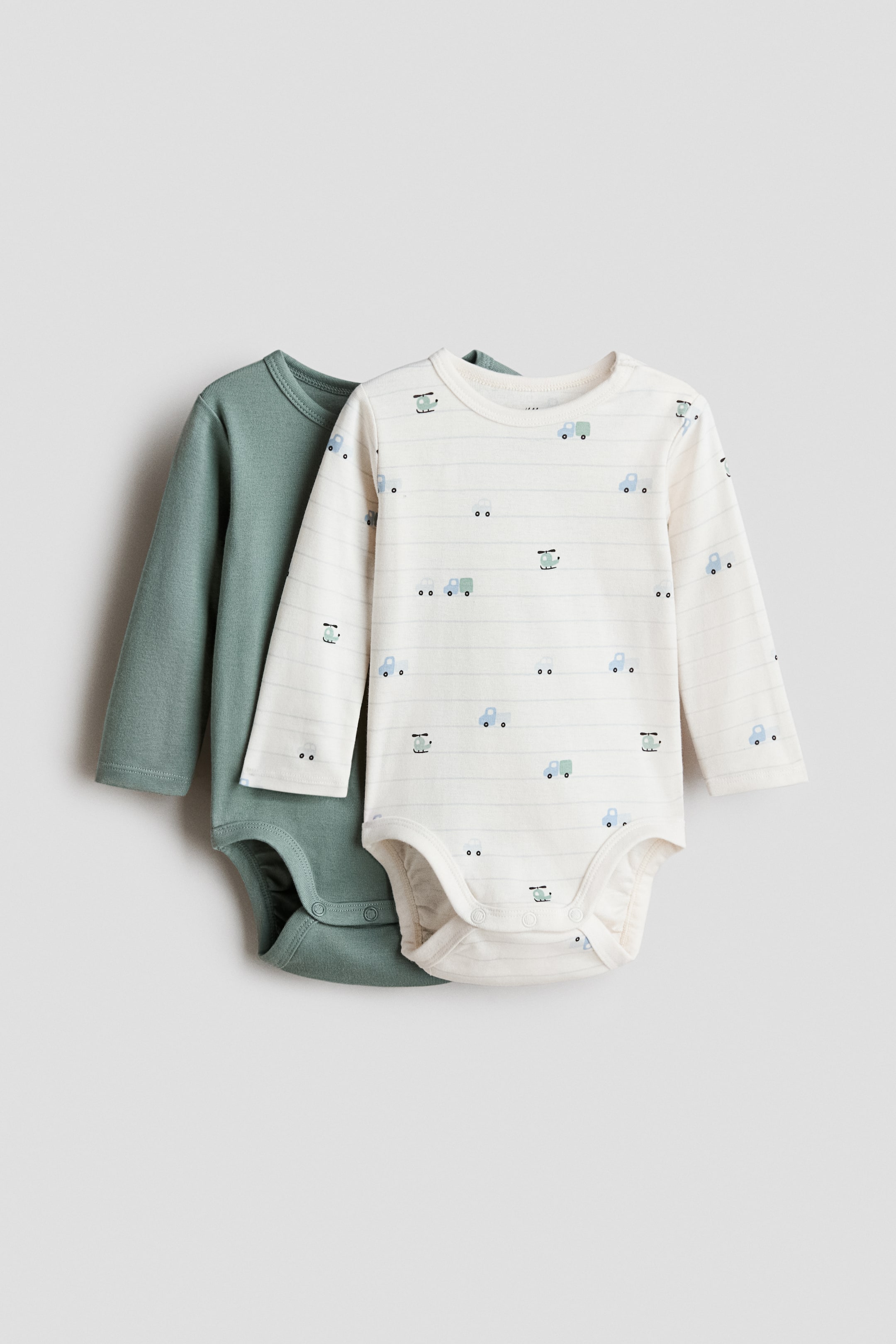 拡大画像を表示: フロントを向いているスーパーソフトなコットンジャージーの長袖ボディスーツのペアが表示されています。1つのボディスーツは無地のセージグリーンで、もう1つはオフホワイトで細い灰色の水平ストライプと小さな青いトラックと緑のヘリコプターのパターンが繰り返し描かれています。どちらのボディスーツもクルーネック、片方の肩に隠しスナップボタン、股間にスナップボタン、脚回りにギャザー縫い目があります。