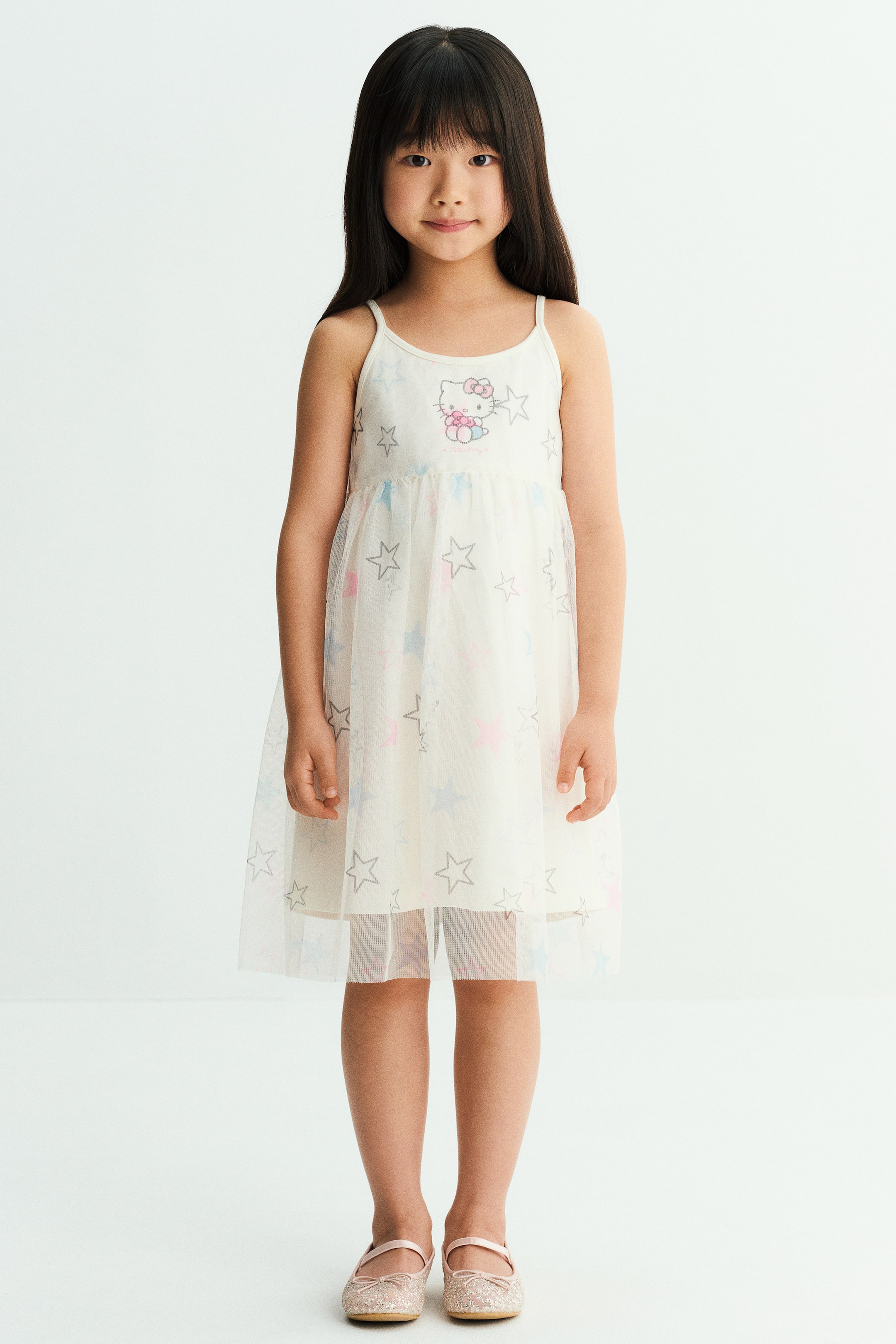 View larger image: A white jersey dress with narrow shoulder straps and a round neckline is worn by the model. The dress features a printed cartoon character on the chest and a flared skirt with a sheer tulle overlay decorated with a multicoloured star print.