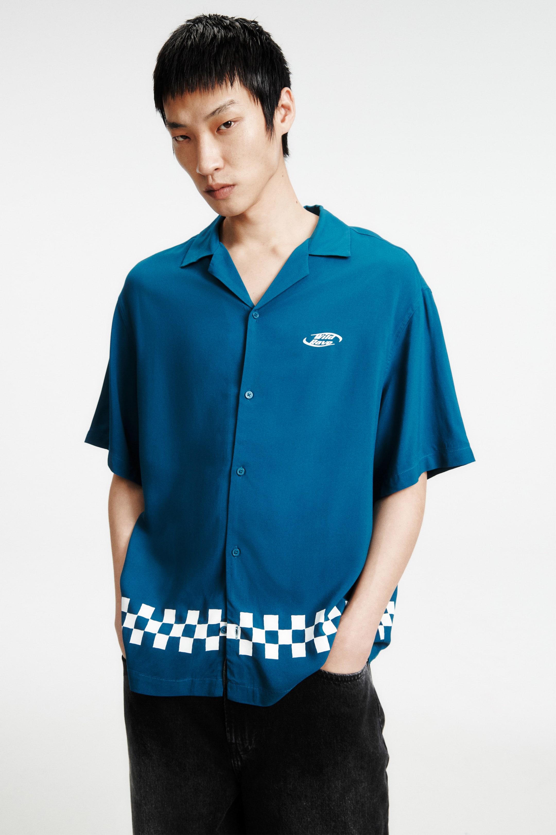 拡大画像を表示: モデルが着用しているのは、リゾートカラーとフレンチフロントを備えた半袖のティールシャツです。このゆったりとしたフィットのシャツには、胸元に白い刺繍のロゴがあり、ストレートカットの裾には白いチェックのバンドが施されています。モデルは、ダークなトラウザーズのポケットに手を入れています。