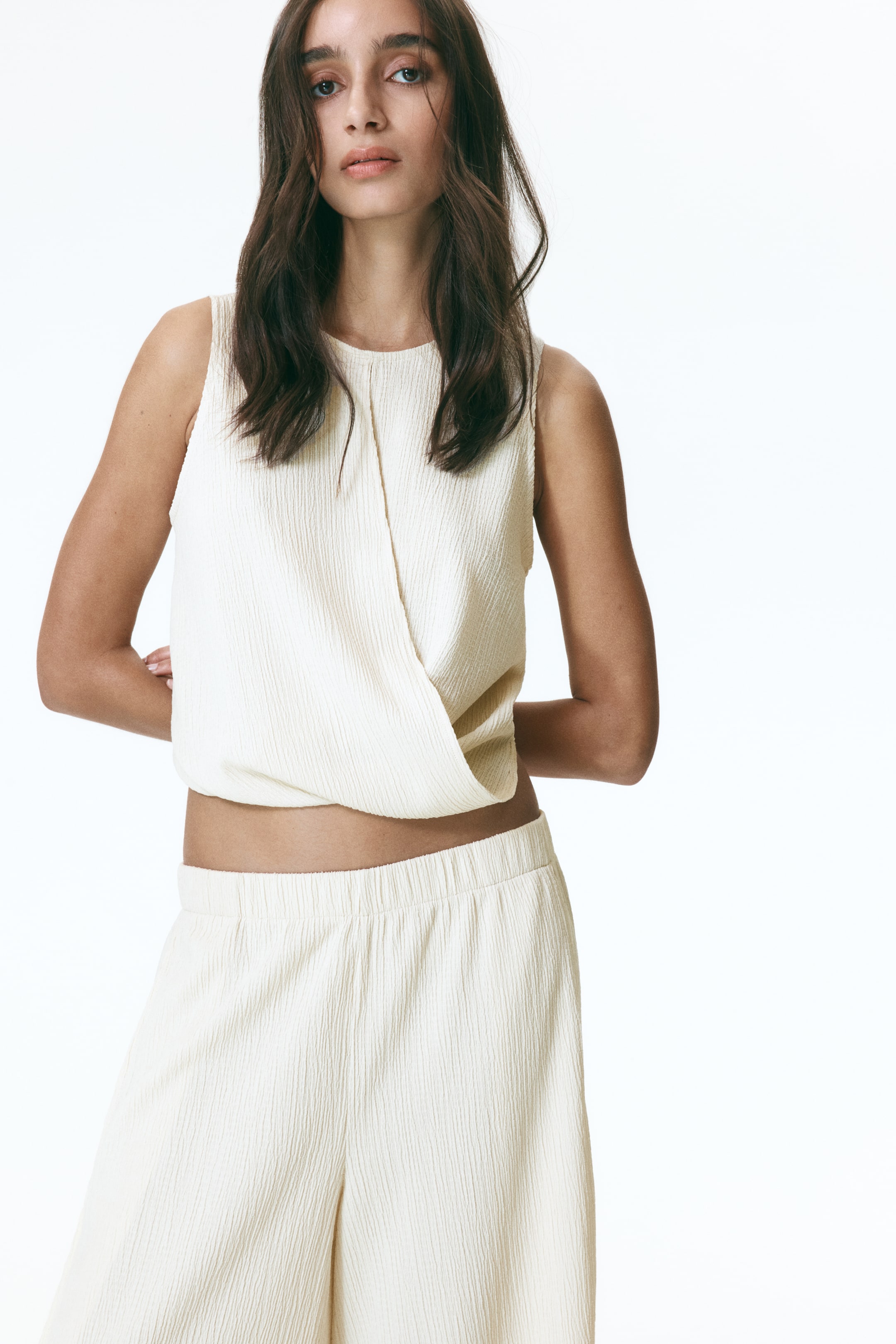 Agrandir l'image: Un ensemble deux pièces texturé crème est présenté, comprenant un haut sans manches avec un col rond et un devant drapé et superposé. Un pantalon à jambes larges assorti avec une taille élastiquée complète l'ensemble.