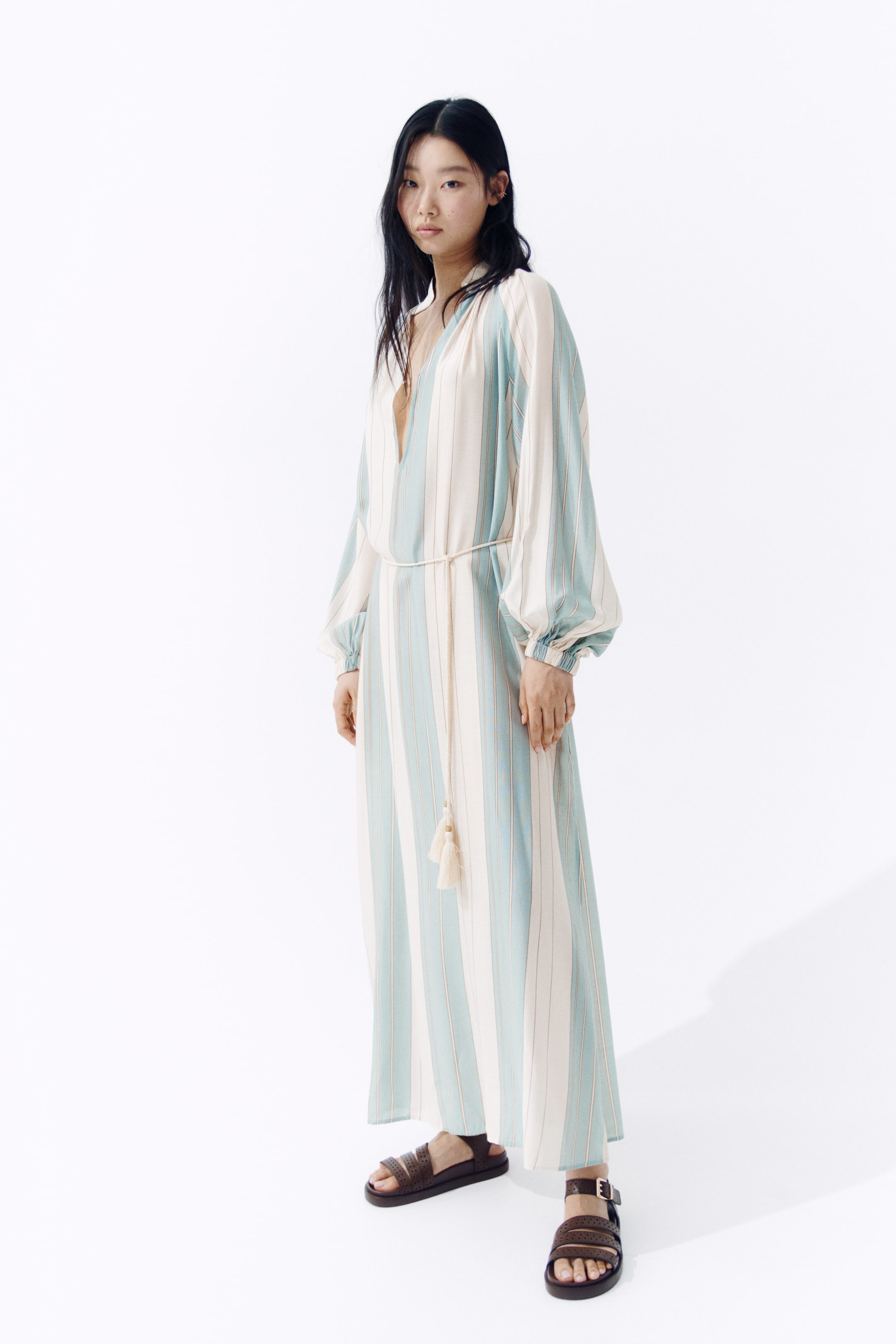 View larger image: A long, oversized tunic dress adorned with light blue and off-white vertical stripes is showcased by the model. It features a stand collar with gathers, a deep V-shaped opening, long balloon sleeves with elasticated cuffs, a narrow tasselled tie belt cinching the waist, and high slits visible at each side of the hem.