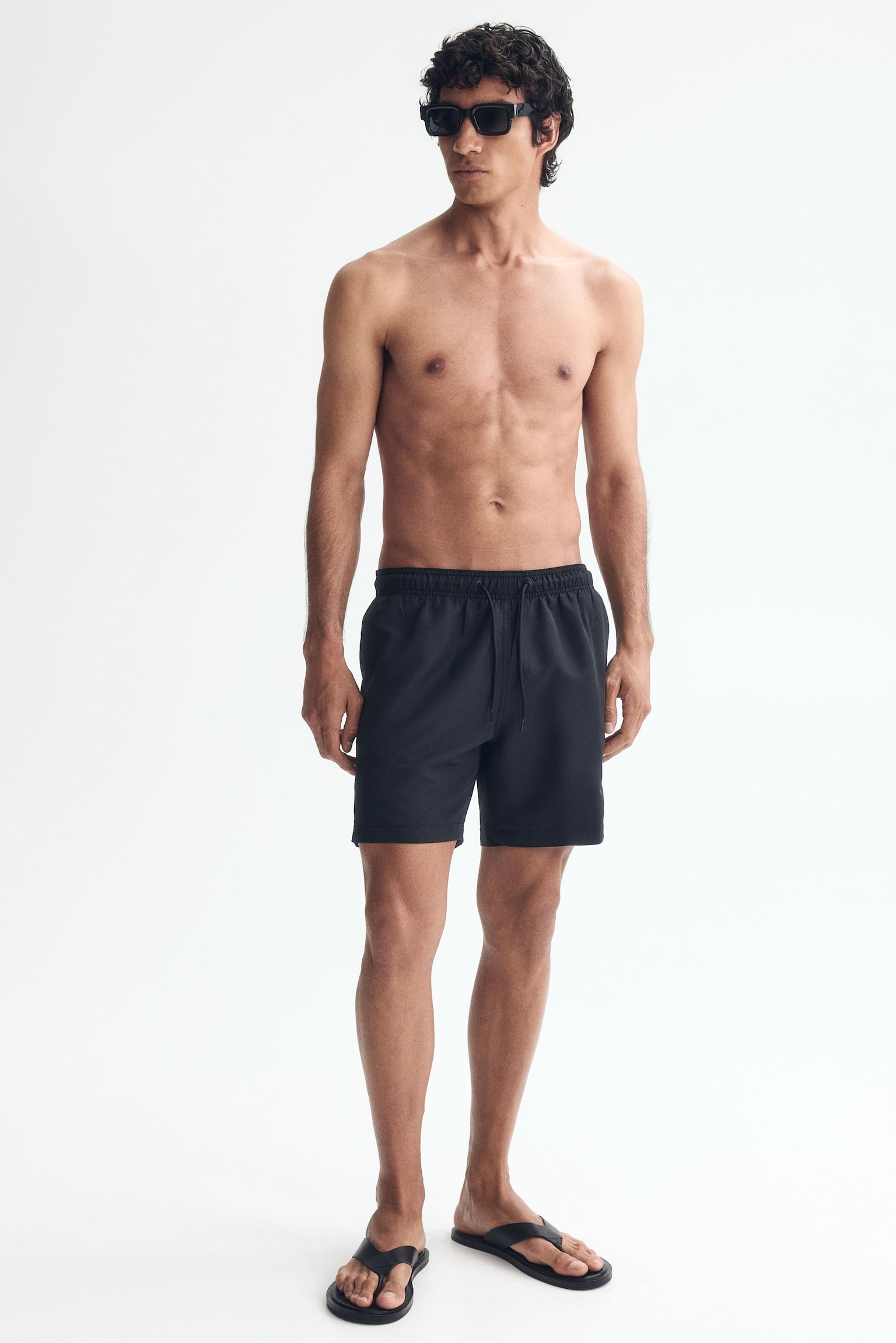 Black shorts are featuring a smooth fabric and an elasticated waistband with a central drawstring tie. They are displaying subtle side seam pockets and small side slits at the hem, presented from a front view. - 1