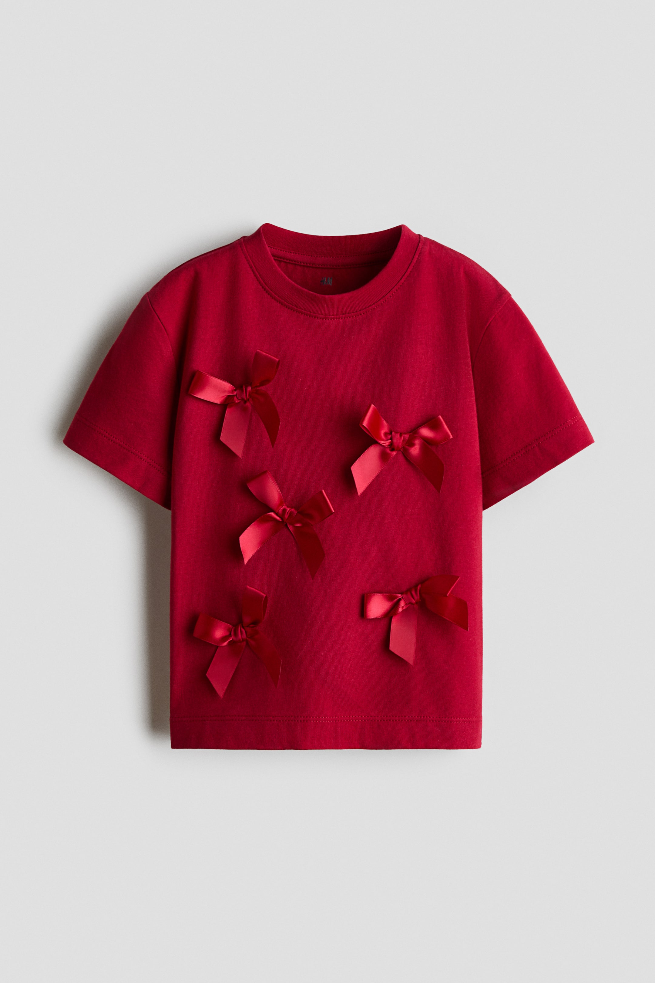 View larger image: A red short-sleeved t-shirt is facing forward, featuring a crew neck and multiple red satin bows scattered across its front surface.