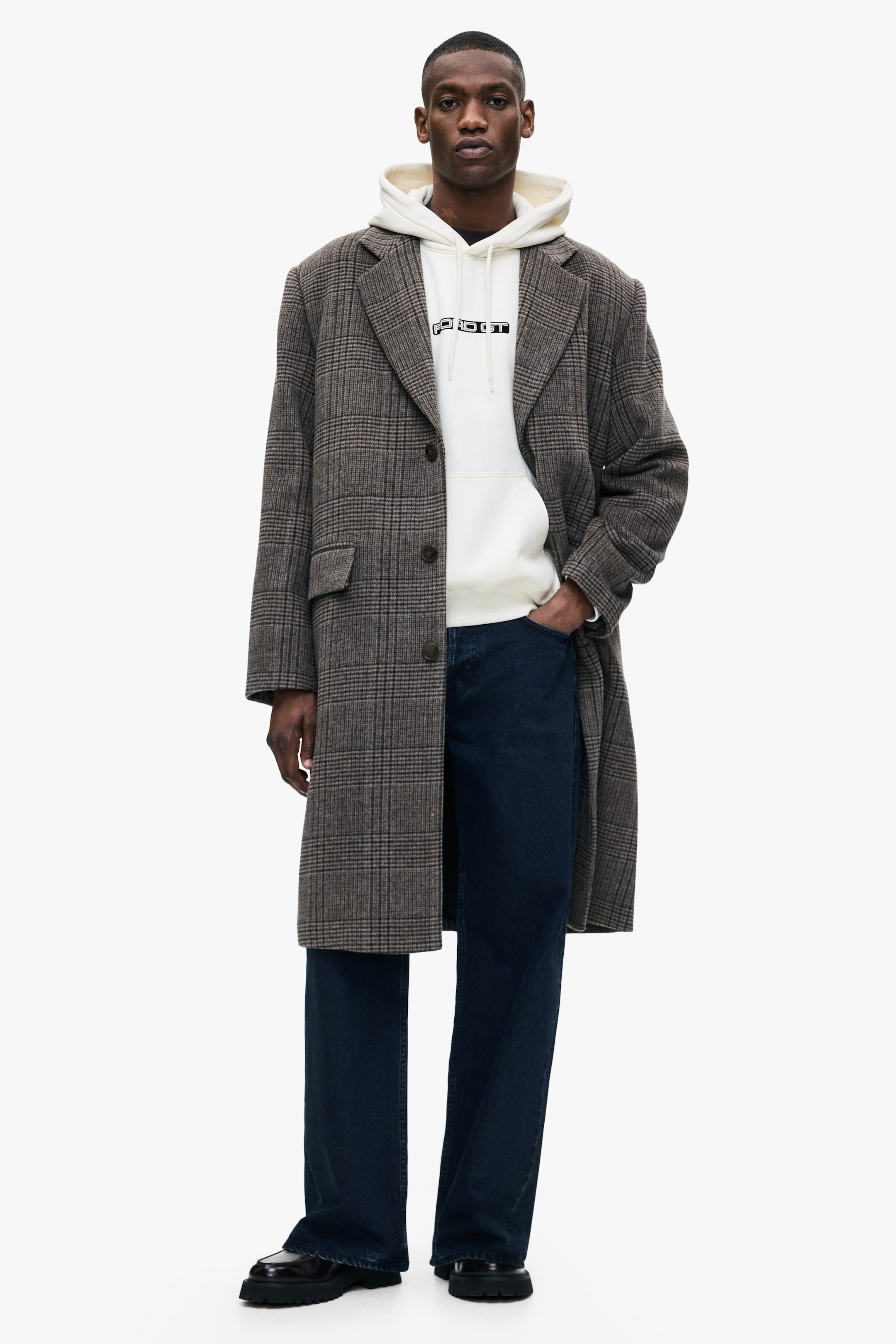 Größeres Bild anzeigen: Ein langer Mantel mit einem braunen und grauen Karomuster, breiten Revers und drei Knöpfen wird offen getragen. Das Model trägt ihn in Lagen über einem cremefarbenen Hoodie mit Grafikdruck, dunkelblauen weiten Jeans und schwarzen groben Loafers.