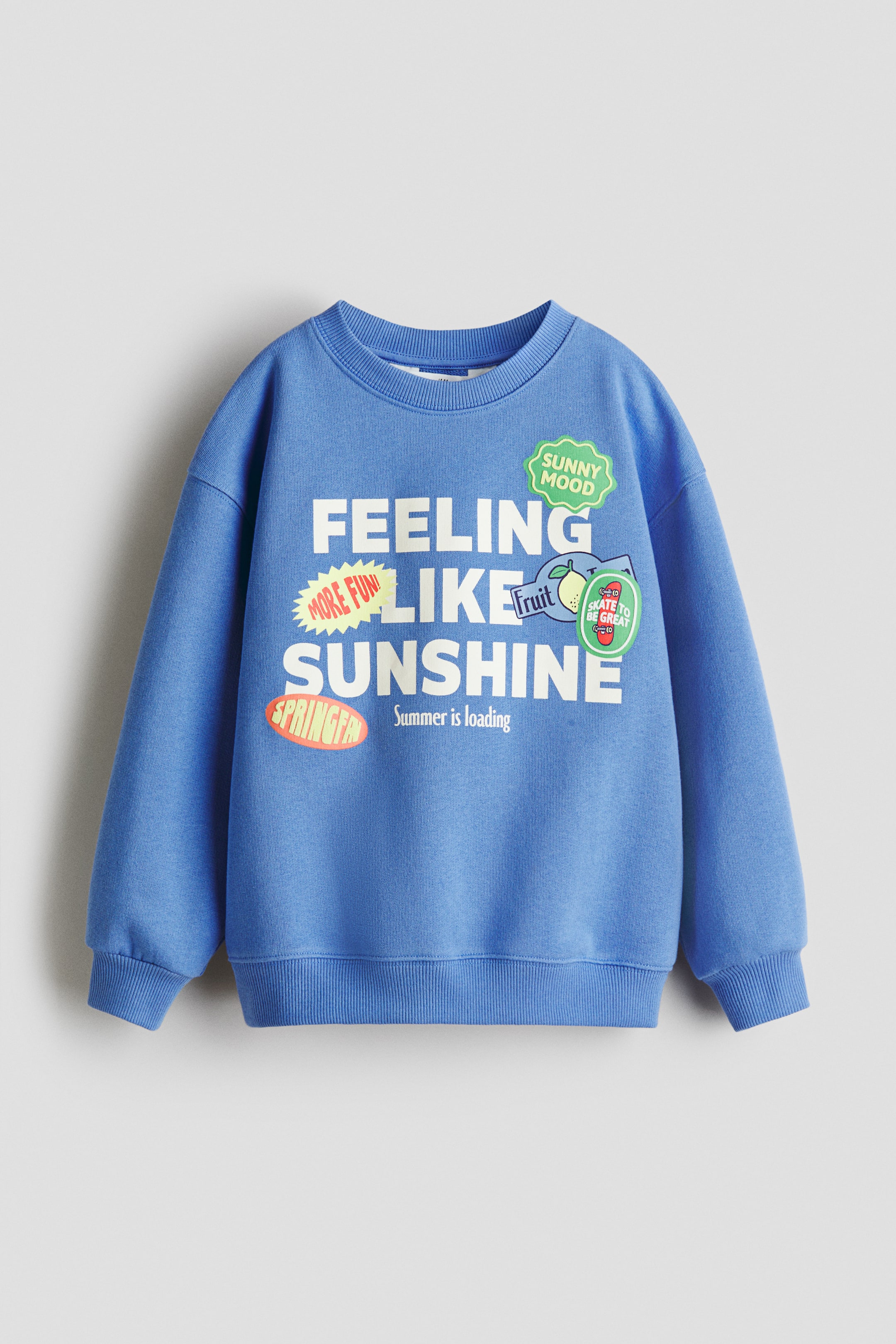 Grotere afbeelding bekijken: Een loszittende blauwe sweatshirt wordt gepresenteerd aan de VOORKANT, met een grote grafische print op de borst met witte tekst 'FEELING LIKE SUNSHINE' en 'Summer is loading', geaccentueerd door kleurrijke patches. Het heeft een ronde halslijn met ribboord, verlaagde schouders, lange mouwen met geribde manchetten, en een geribde zoom.