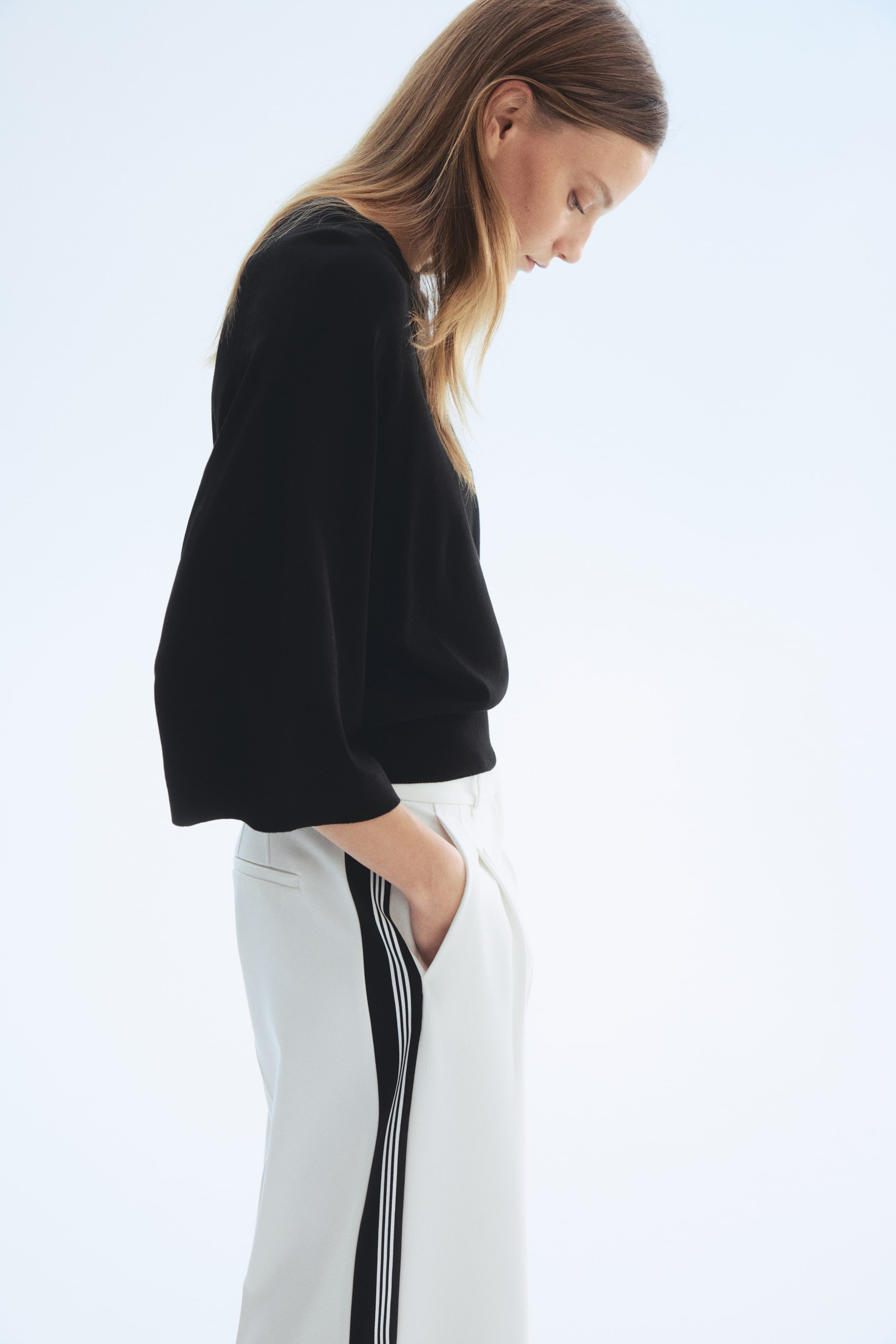 Zobacz większy obraz: Czarna bluzka z szerokimi, trzy-czwartymi rękawami i prążkowanym dołem. Nosi się ją z białymi spodniami z czarnym paskiem bocznym i trzema cienkimi białymi liniami, z ręką spoczywającą w kieszeni.