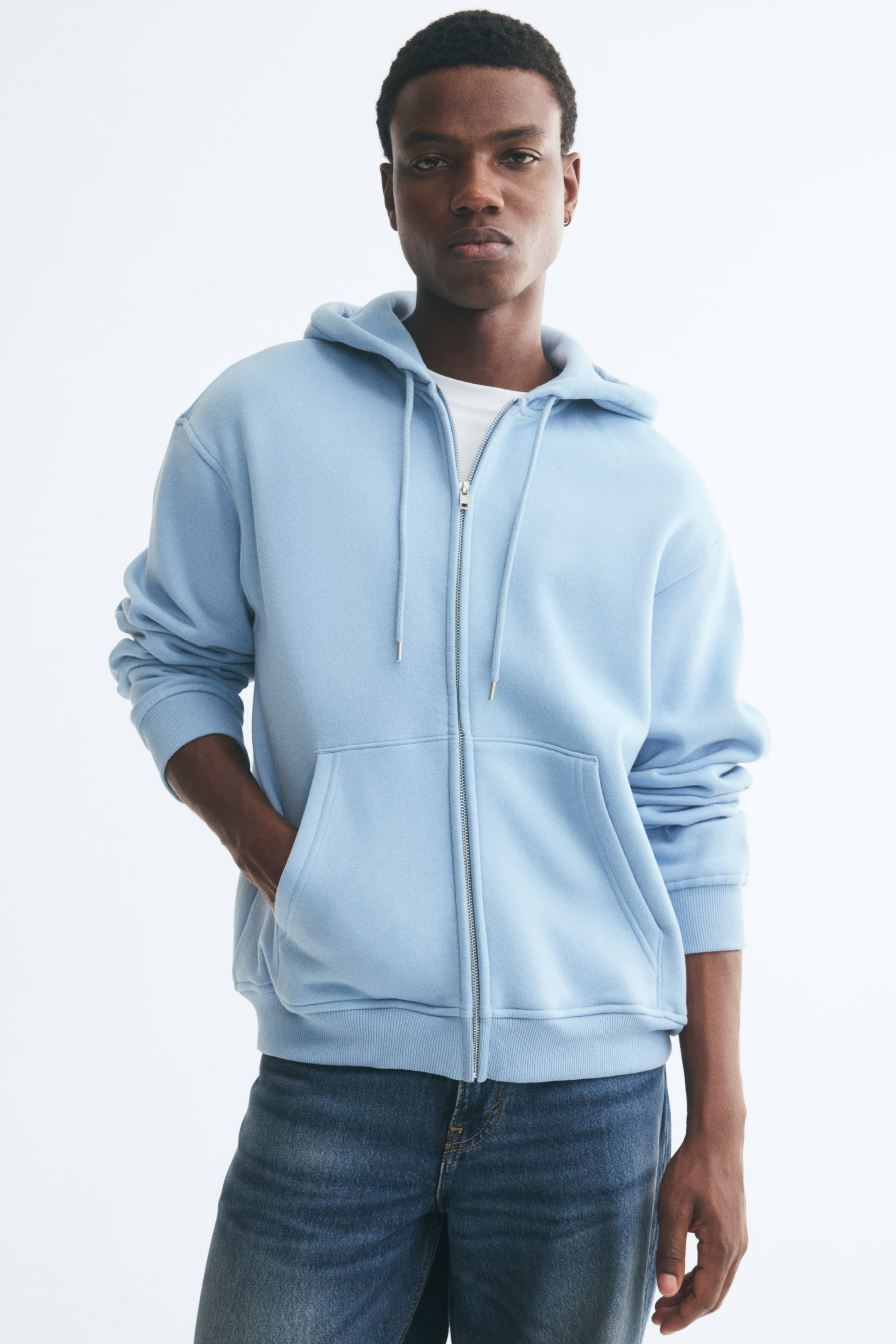View larger image: A light blue zip-up hoodie is worn by the model over a white T-shirt and blue jeans, with one hand placed in a front pocket. The hoodie features a drawstring hood, dropped shoulders, a front metal zip, and ribbed trims at the cuffs and hem.