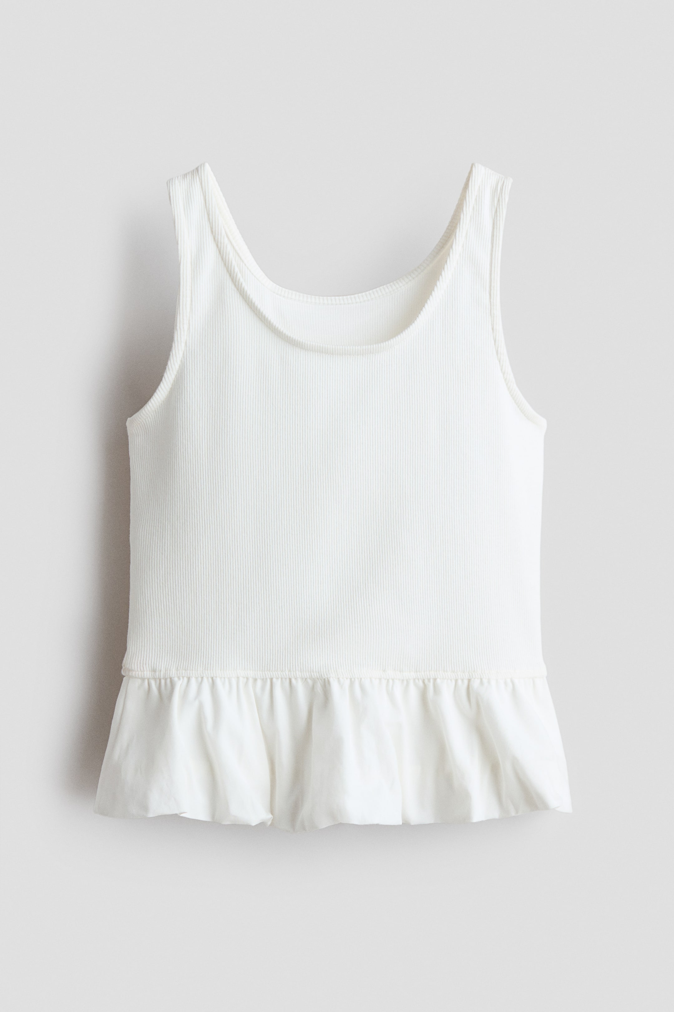 Größeres Bild ansehen: Das weiße, nach vorne gerichtete Oberteil verfügt über ein geripptes Tanktop-Oberteil mit breiten Trägern und einem Rundhalsausschnitt. Es geht in einen gerafften Schößchen-Saum über, der in einem glatteren Stoff erscheint.