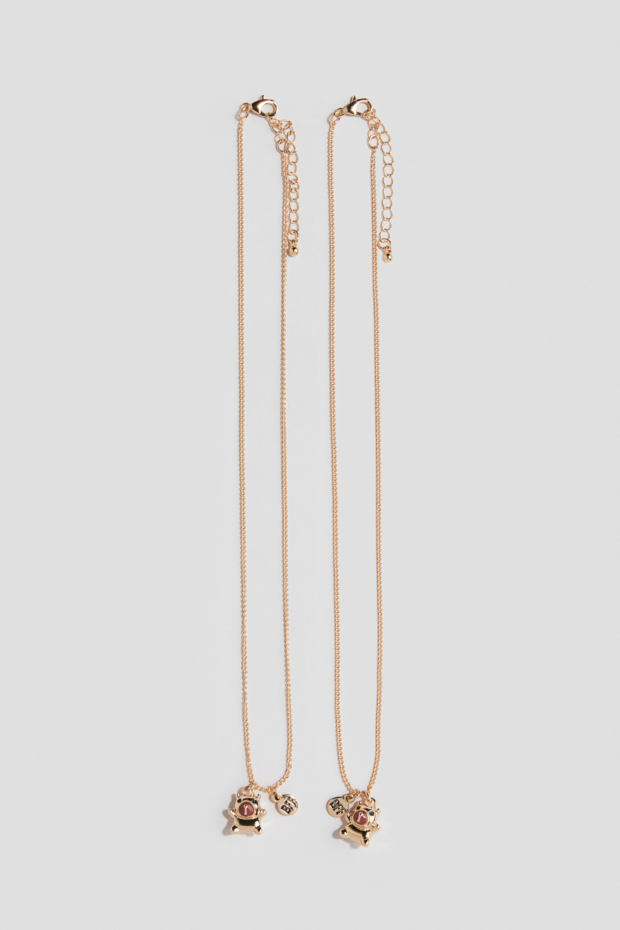 Ver imagen más grande: Dos collares a juego de tono dorado reposan en vertical, con finas cadenas de eslabones tipo cable, cierres de mosquetón y cadenas extensibles ajustables. Cada collar muestra un pequeño colgante frontal con forma de oso que lleva un casco rosa, junto a un pequeño charm circular grabado con las letras "BFF".