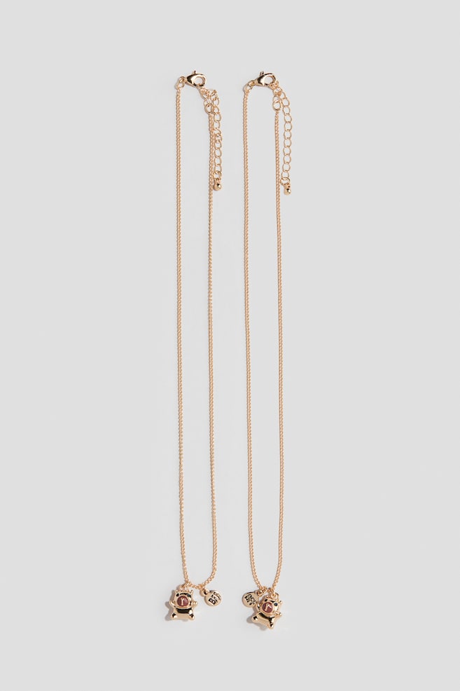 Two matching gold-toned necklaces are lying vertically, featuring fine cable chains with lobster claw clasps and adjustable extender chains. Each necklace is displaying a small, front-facing bear-shaped pendant wearing a pink helmet alongside a small circular charm engraved with the letters "BFF". - 1