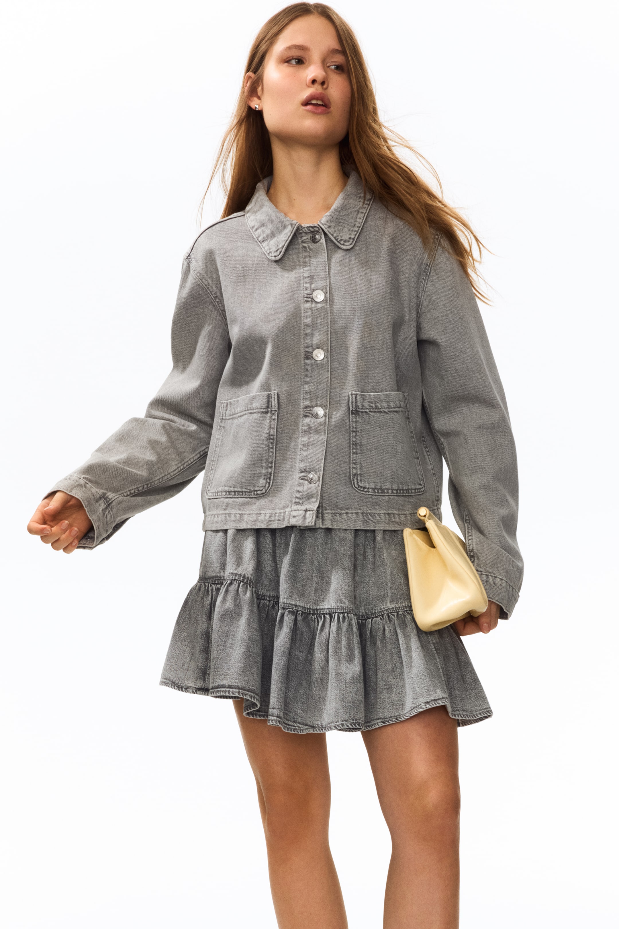 Büyük resmi görüntüle: Model, yaka, önden düğmeli, düğmeli manşetler ve ön cep detayları olan kısa, gri, dar kesim pamuklu denim ceket giymektedir. Model ayrıca kat kat denim etek giymekte ve açık renkli bir çanta tutmaktadır.