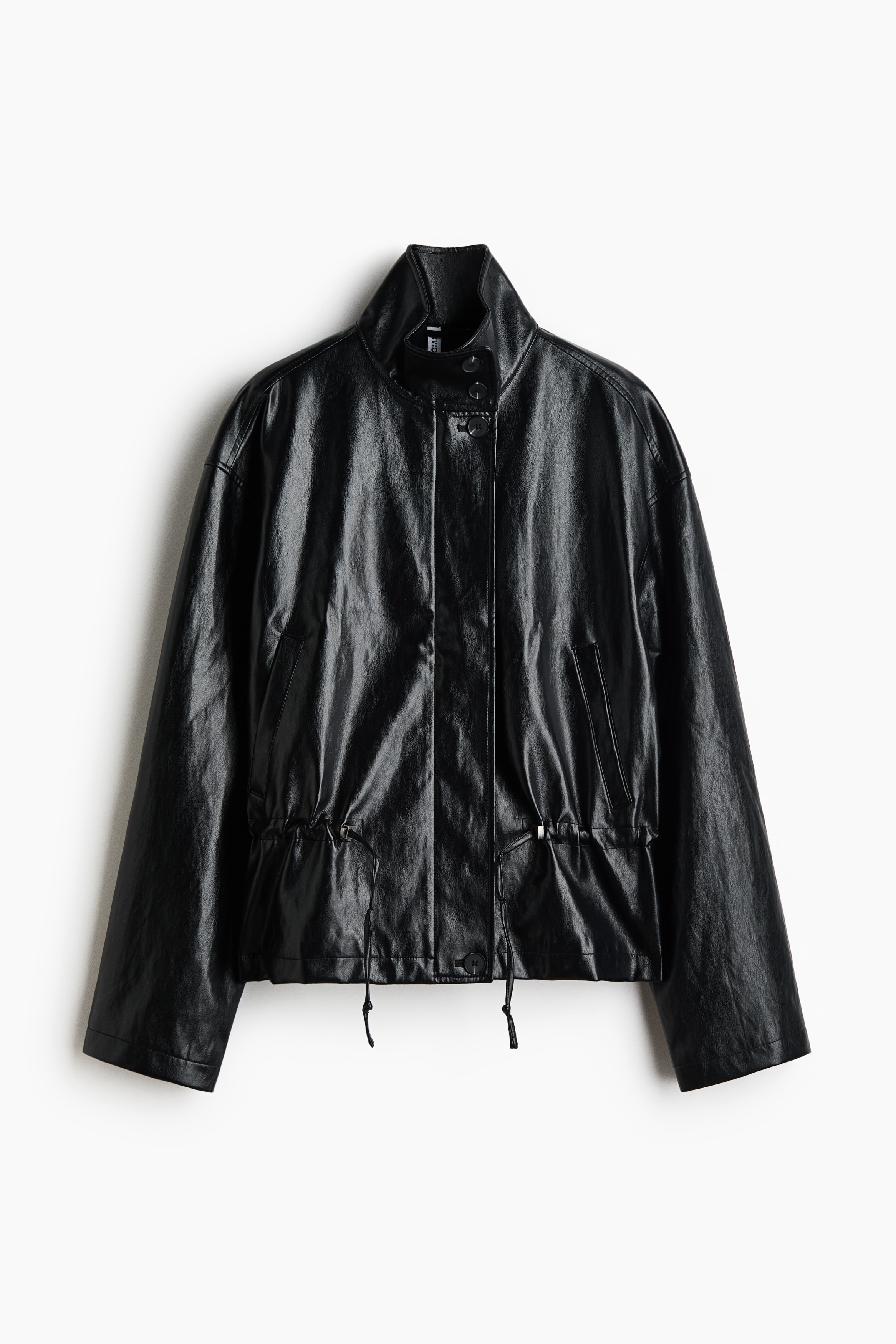 Das Model trÃ¤gt eine Oversize-Jacke aus glÃ¤nzendem, schwarz beschichtetem Stoff und hat eine Hand in der Leistentasche vorne. Die Jacke verfÃ¼gt Ã¼ber einen Stehkragen, Ã¼berschnittene Schultern und einen gerafften Kordelzug in der Taille und ist mit blauen Jeans kombiniert.