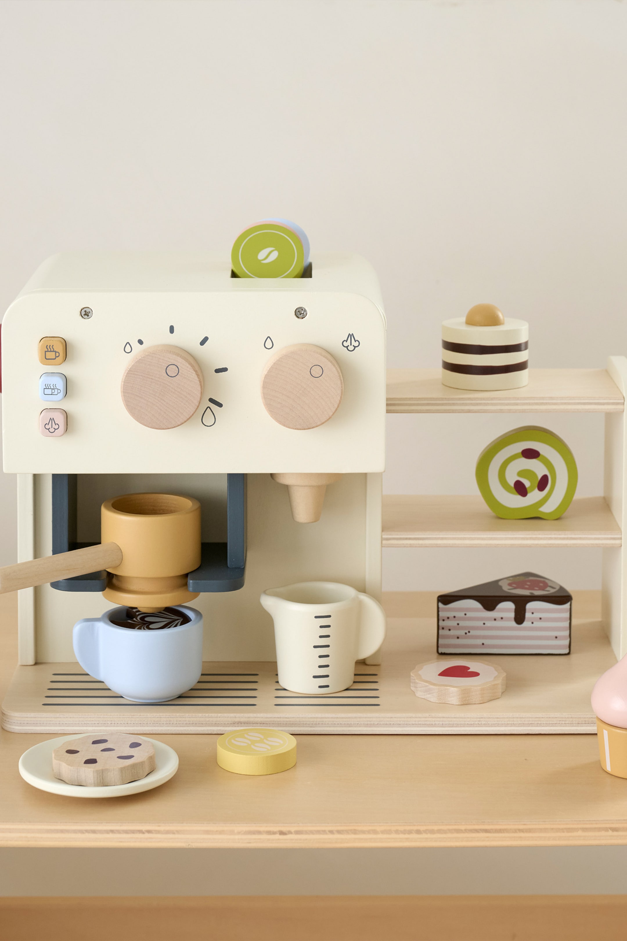 Grotere afbeelding bekijken: Een houten speelgoed koffiemachine speelset met een crèmekleurige machine met twee houten bedieningsknoppen, drie vierkante knoppen en een portafilter die in een lichtblauw kopje met latte art schenkt. De set bevat ook een zijplank met verschillende houten speelvoedselitems zoals taarten en koekjes, samen met een maatbeker, een koekje op een bord en een schijfje citroen.