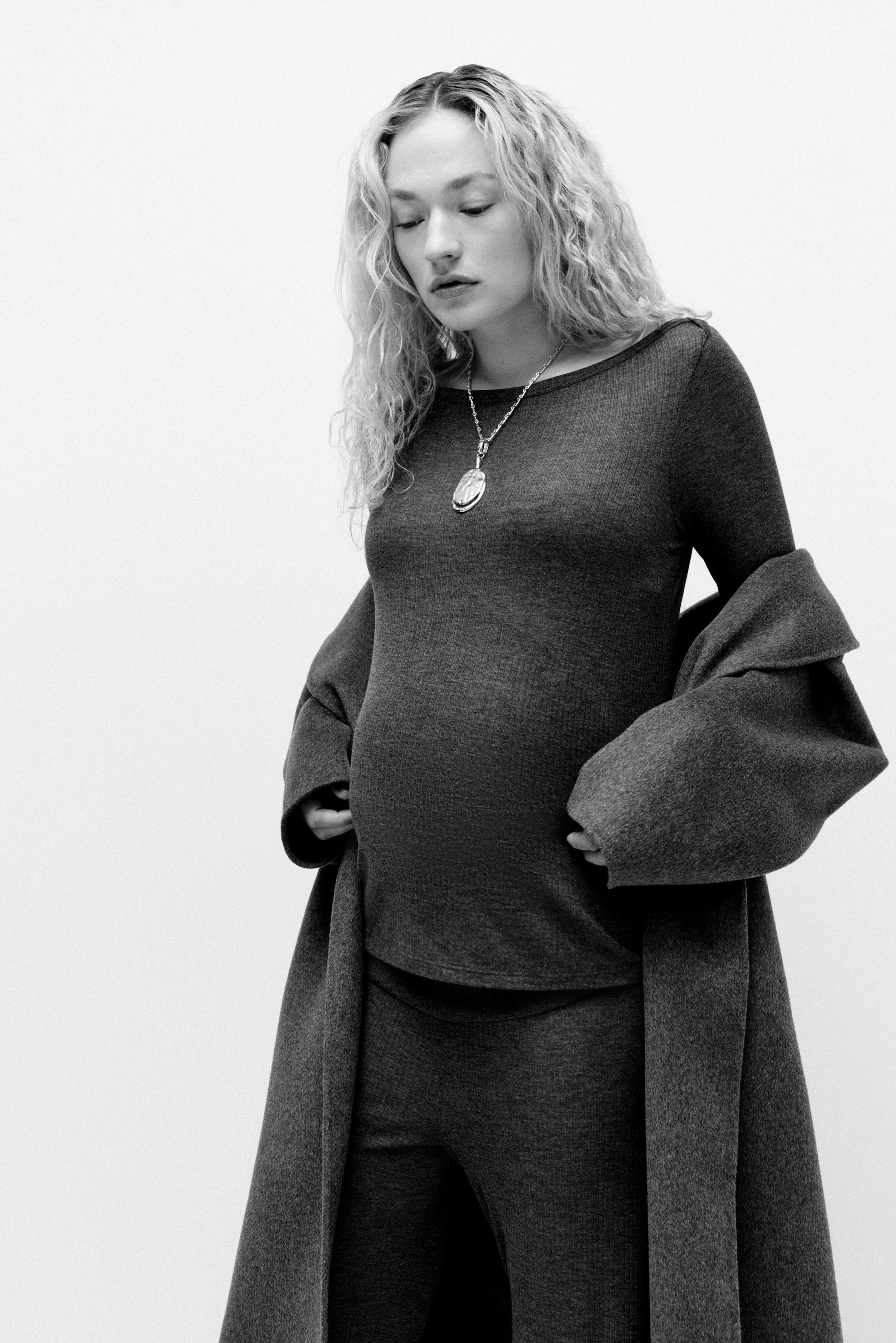 Vis større bilde: Et sett i mørke toner, bestående av en langermet, ribbestrikket jersey-topp med båtutringning og rette bukser med bredt midjepanel, bæres av modellen. Modellen plasserer hendene på magen og interagerer med det brede midjepanelet, mens en frakk er drapert over skuldrene hennes.