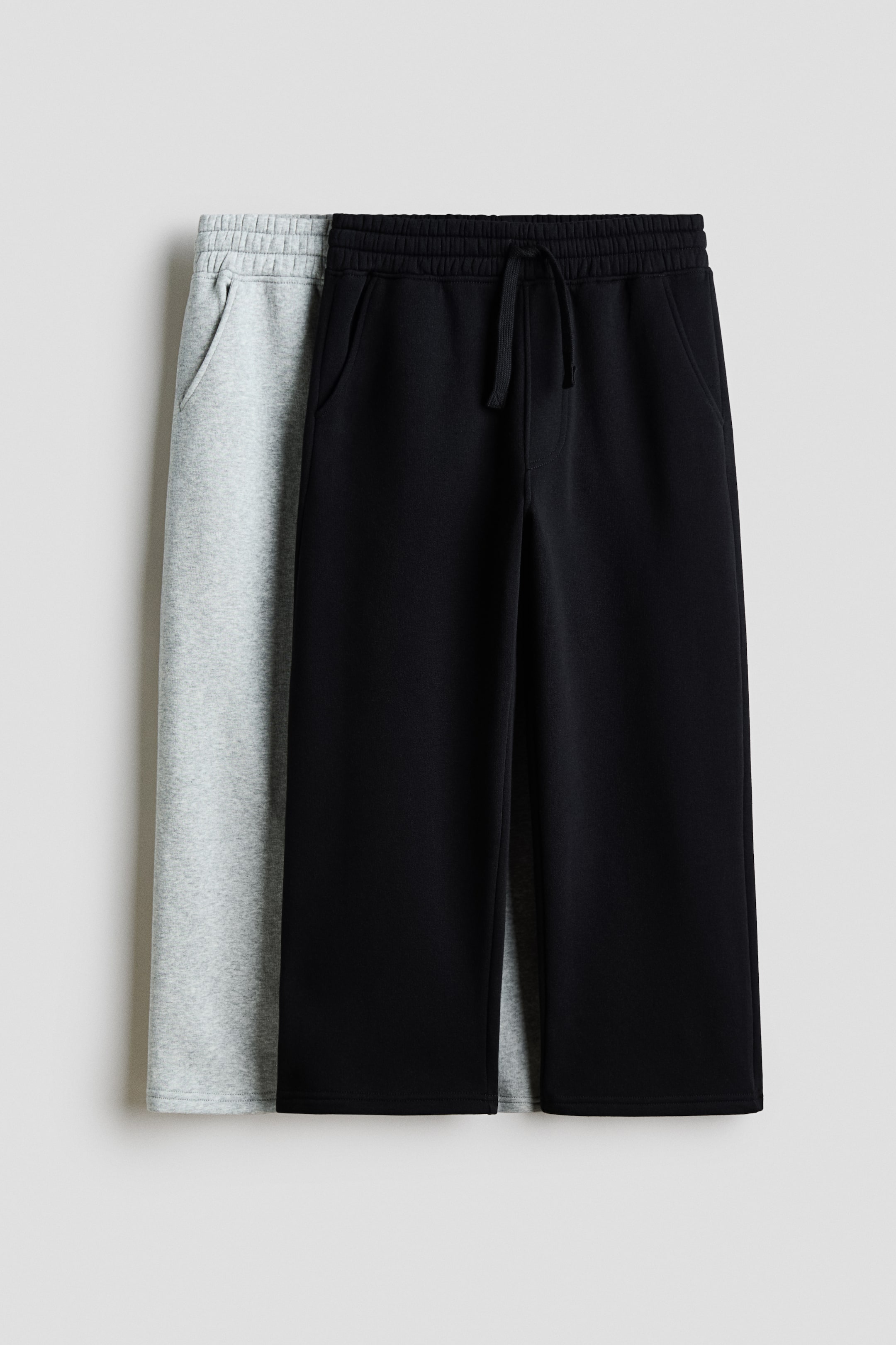 View larger image: Two pairs of wide-leg fleece joggers are hanging in a front view, with a black pair layered directly in front of a light grey pair. Both pairs feature an elasticated waistband with a drawstring, diagonal side pockets, a fake fly, and a relaxed, wide-leg cut.