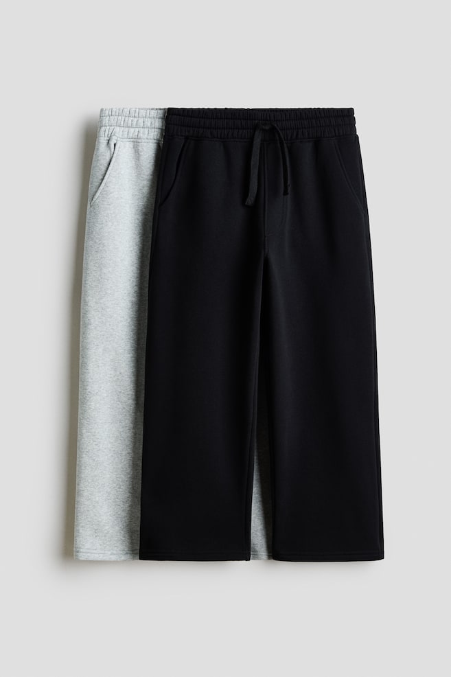 Zwei Paar Fleece-Jogginghosen mit weitem Bein hÃ¤ngen in einer Frontansicht, wobei ein schwarzes Paar direkt vor einem hellgrauen Paar hÃ¤ngt. Beide Paare verfÃ¼gen Ã¼ber einen elastischen Bund mit Kordelzug, schrÃ¤ge Seitentaschen, einen Fake-ReiÃverschluss und einen lockeren, weit geschnittenen Beinverlauf. - 1