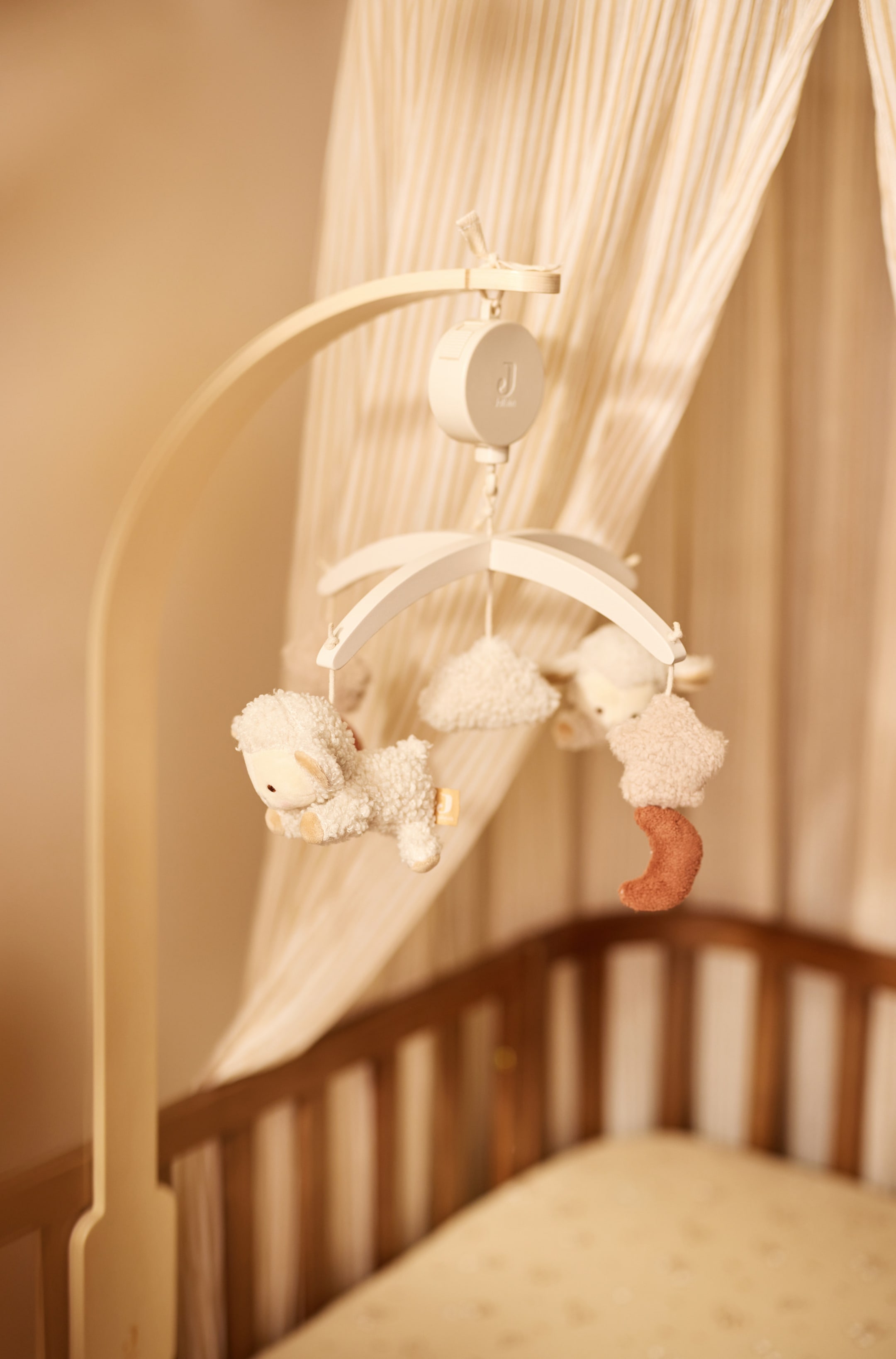 Grotere afbeelding bekijken: Een baby mobiel hangt aan een gebogen houten arm, met een centraal mechanisch onderdeel en een witte dwarsstangophanging. Zachte, getextureerde figuurtjes, waaronder twee kleine schaapjes, een wolk, een ster en een roodbruine maansikkel, hangen aan de dwarsstang.